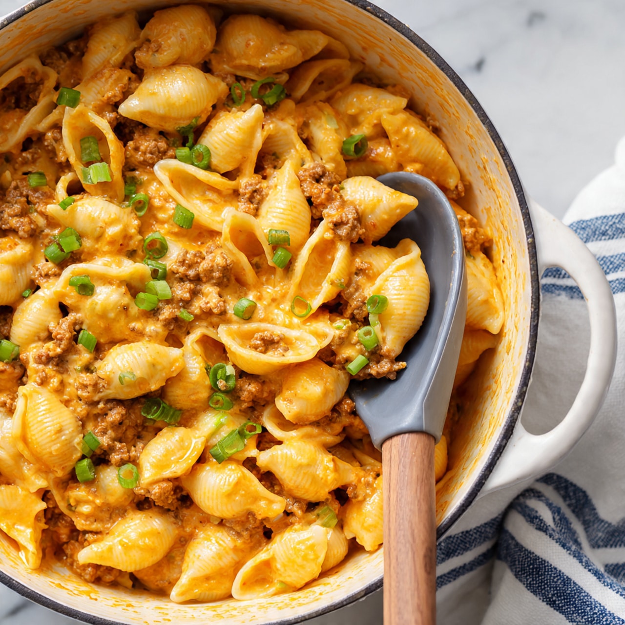 A close-up view of a large pot filled with creamy orange cheese pasta shells mixed with browned ground meat, scattered with bright green chopped scallions on top. The pasta shells are coated evenly in a smooth, thick cheese sauce with visible small pieces of meat intertwined. A gray spoon with a wooden handle is scooping some of the pasta from the pot, showing the soft texture of the shells and sauce. The background features a white marbled surface with a white and navy striped cloth partially visible. photo taken with an iphone --ar 4:5 --v 7