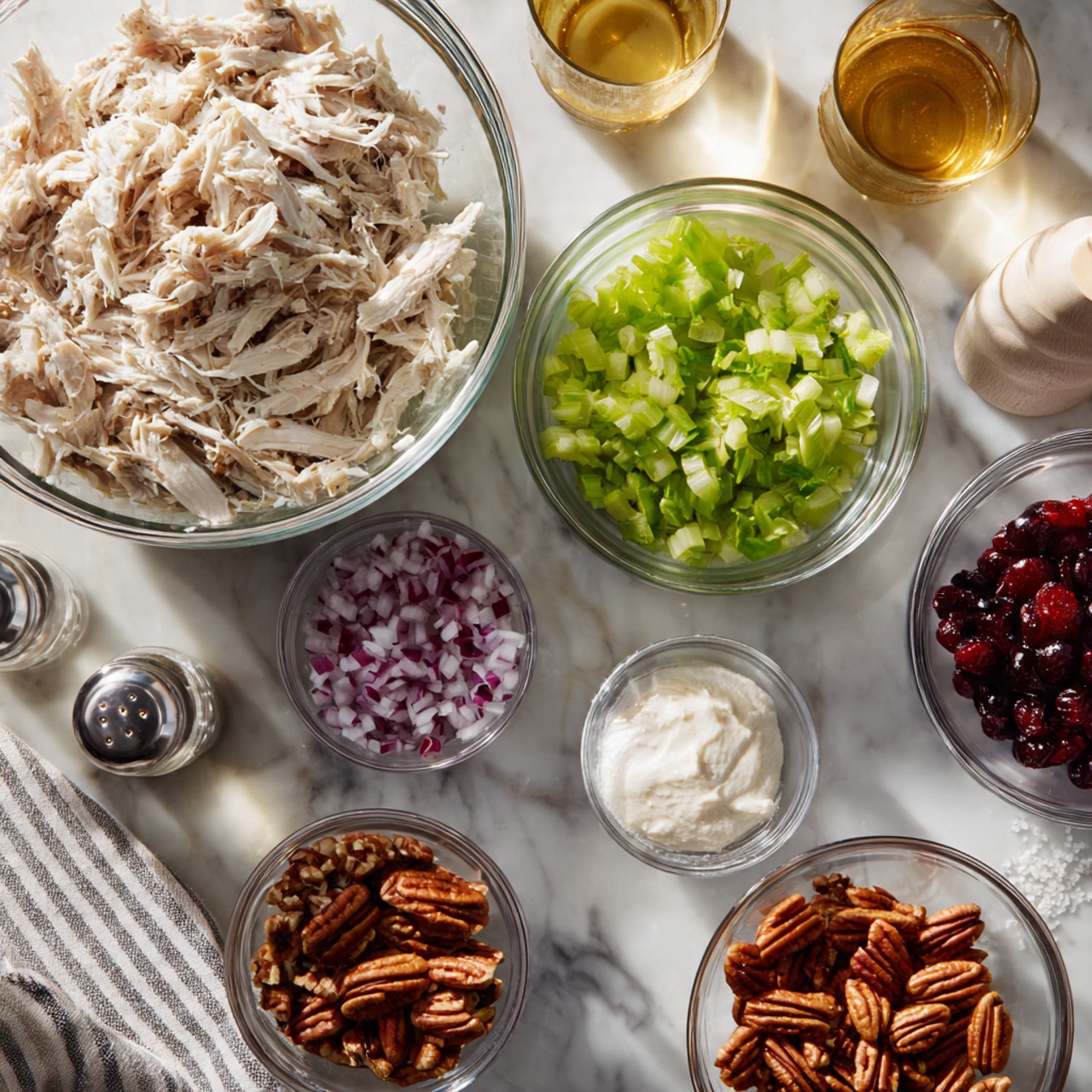The image shows a white marbled surface with several clear glass bowls containing different ingredients arranged neatly. In the top left, there is a large bowl filled with shredded white meat. Surrounding it are smaller bowls: one with chopped light green celery, one with red dried cranberries, one with finely chopped purple onions, one with whole pecans, a small bowl with a white creamy substance, and another small bowl with a thick white mixture. There are also salt and pepper shakers, and a small glass with a golden liquid. A white cloth with gray stripes is partly visible at the bottom left. The lighting is bright and natural, enhancing the crisp and clean appearance. photo taken with an iphone --ar 4:5 --v 7