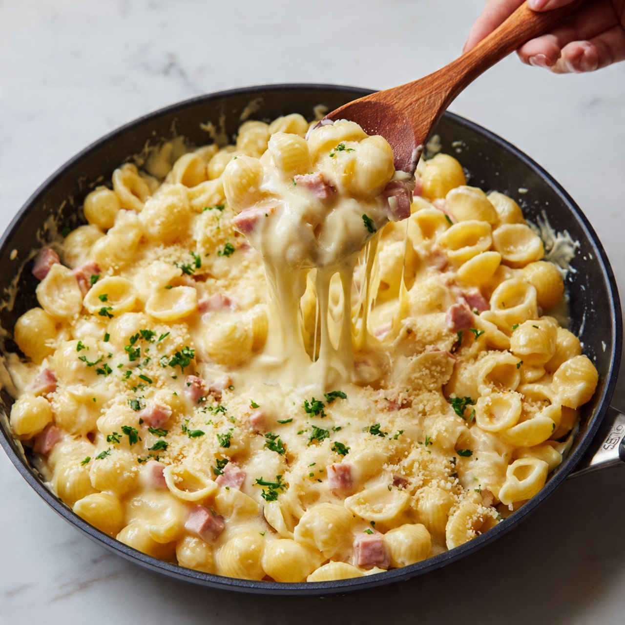 A close-up image of a creamy macaroni and cheese dish with small pasta shells covered in melted yellow cheese. There are small pieces of pink ham mixed into the pasta, and fresh green chopped herbs sprinkled on top. In the center, a wooden spoon held by a woman's hand lifts a portion of the cheesy pasta, showing the thick, creamy cheese sauce stretching slightly. The dish is served in a black pan placed on a white marbled surface. photo taken with an iphone --ar 4:5 --v 7