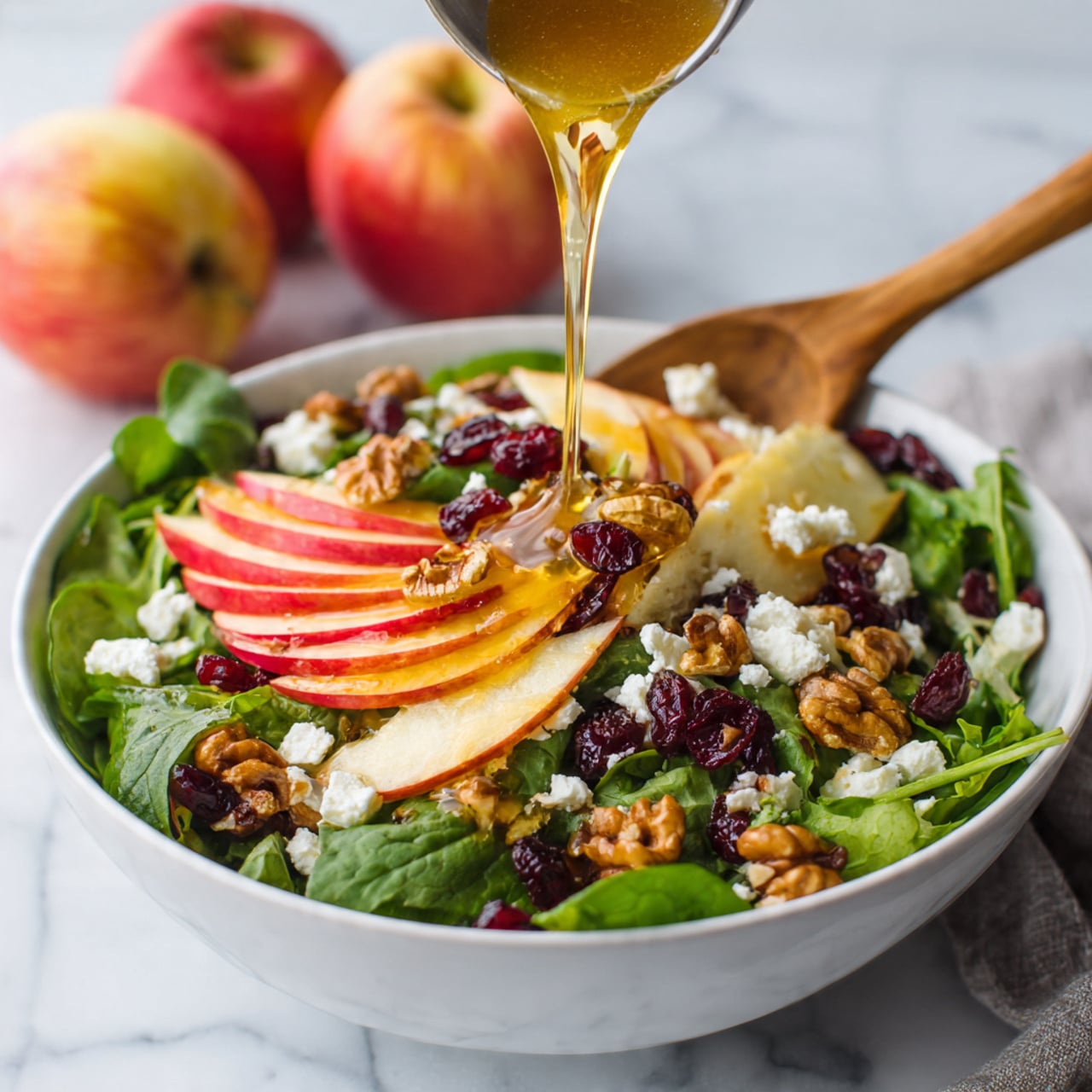 A white bowl filled with a fresh salad placed on a white marbled texture. The salad has a base layer of mixed green lettuce and spinach leaves. On top of the greens are several thin slices of red and yellow apple, spread around the edges of the bowl. Crumbled white cheese is scattered evenly over the salad, along with dried dark red cranberries and golden-brown walnuts. A wooden spoon rests inside the bowl, and a golden liquid dressing is being poured gently over the top. Three whole apples with red and yellow skin are visible in the blurred background. Photo taken with an iphone --ar 4:5 --v 7