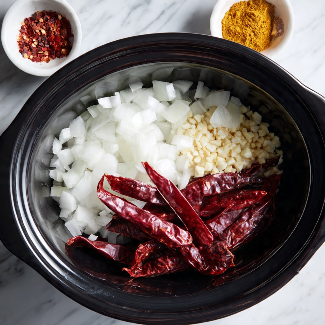Inside a black slow cooker, there are three main layers of ingredients: at the bottom, chopped white onions with a soft texture; next to them, several whole dried red chili peppers with a rough, wrinkled surface; above them, tiny minced garlic pieces scattered unevenly. The slow cooker is set against a white marbled surface with a small white bowl of yellow spices partially visible to the side. Photo taken with an iphone --ar 4:5 --v 7