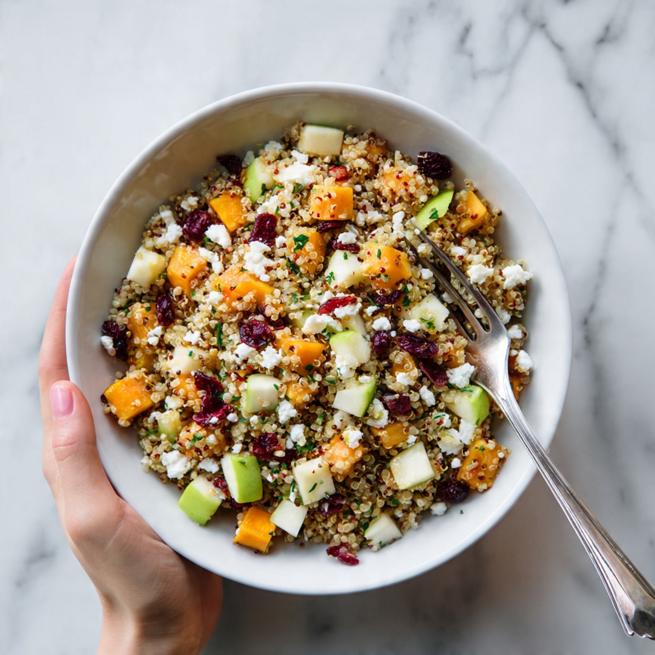 A white bowl filled with a colorful salad showing a mix of small quinoa grains as the base layer, topped with roasted orange cubes of butternut squash, light green diced apple pieces, and small bright red dried cranberries scattered throughout. There are some crumbled white feta cheese bits on top, adding a soft texture. A woman's hand holds a silver fork resting on the side of the bowl. The bowl sits on a white marbled surface. photo taken with an iphone --ar 4:5 --v 7