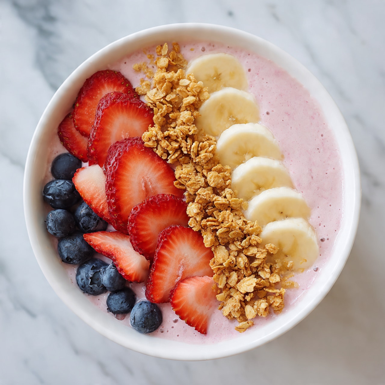 A white bowl filled with a thick, creamy pink smoothie as the main layer, topped on one side with fresh, sliced red strawberries arranged closely together, a cluster of golden brown granola next to the strawberries, a neat row of round, pale yellow banana slices along the bottom edge, and several plump, deep blue blueberries lined up on the left edge of the smoothie, all placed on a white marbled surface photo taken with an iphone --ar 4:5 --v 7