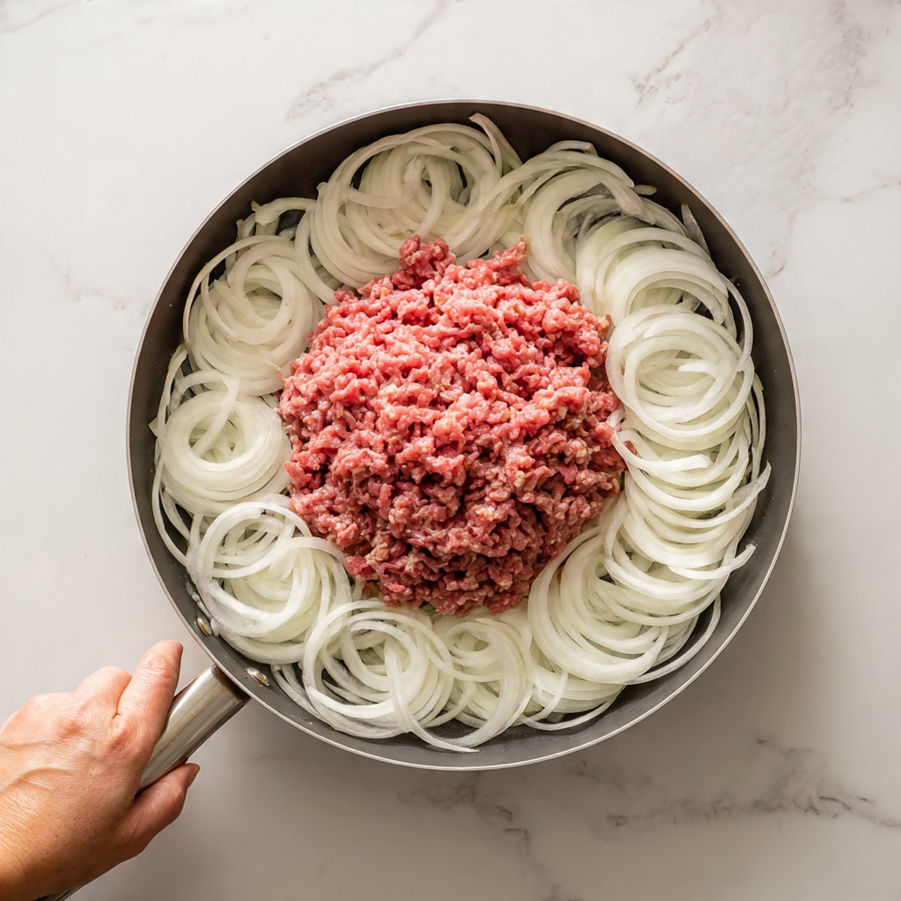 The image shows a round cooking pan with a layer of raw, thinly sliced white onions spread evenly around the edge, forming a ring. In the center of the pan, there is a pile of raw ground beef with a bright red color, slightly uneven in texture. The onions have a soft, translucent look, and the beef looks fresh and moist. The pan sits on a white marbled surface, and there is a woman's hand holding the handle of the pan on the left side. Photo taken with an iphone --ar 4:5 --v 7