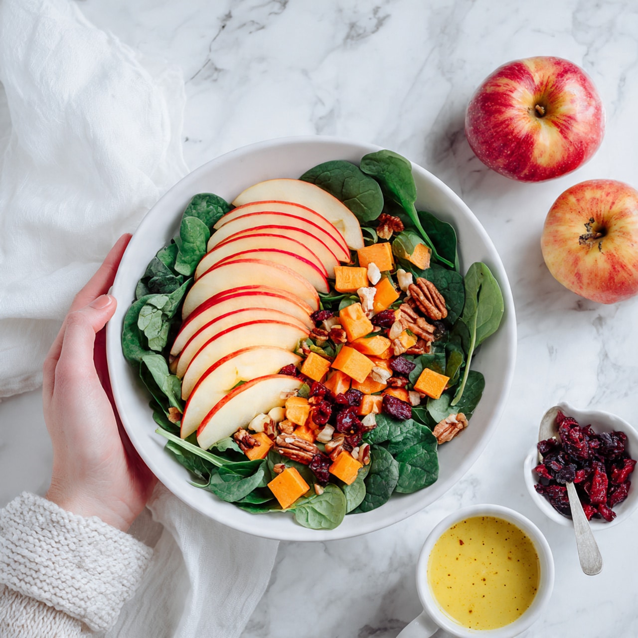 The image shows a white bowl filled with a colorful salad on a white marbled surface. The salad has multiple layers: at the bottom, there are fresh dark green spinach leaves; on top of these are several slices of red and yellow apple arranged in a fan shape on one side. Scattered over the salad are small orange cubes of roasted sweet potato and dark brown nuts. There are also some red dried berries sprinkled around. Nearby, two whole apples sit on the white marbled surface, one darker red and one yellow with red patches. A small white bowl with more dried berries and nuts, and a white cup with yellow dressing and a white spoon, are at the bottom of the image. A woman's hand slightly lifts a white cloth at the top left corner. The overall look is bright and fresh photo taken with an iphone --ar 4:5 --v 7
