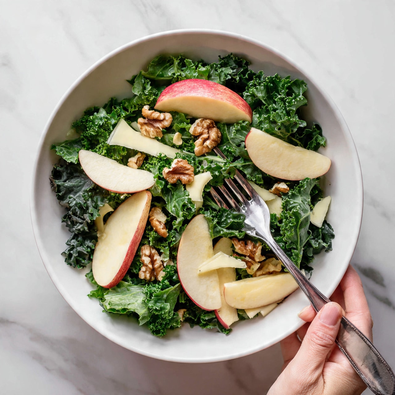 The image shows a fresh salad in a white bowl on a white marbled surface. The salad has several layers starting with dark green kale leaves at the bottom, which have a rough texture. On top of the kale, there are thin, smooth slices of red apple with white flesh, spread evenly around. Mixed in are pale beige slices of cheese with a soft, slightly crumbly texture. Small brown walnut pieces are scattered across the salad, adding a rough and crunchy look. A silver fork rests in the bowl, and a woman's hand is holding the fork, ready to take a bite. The colors are natural and vibrant, making the salad look fresh and healthy. photo taken with an iphone --ar 4:5 --v 7