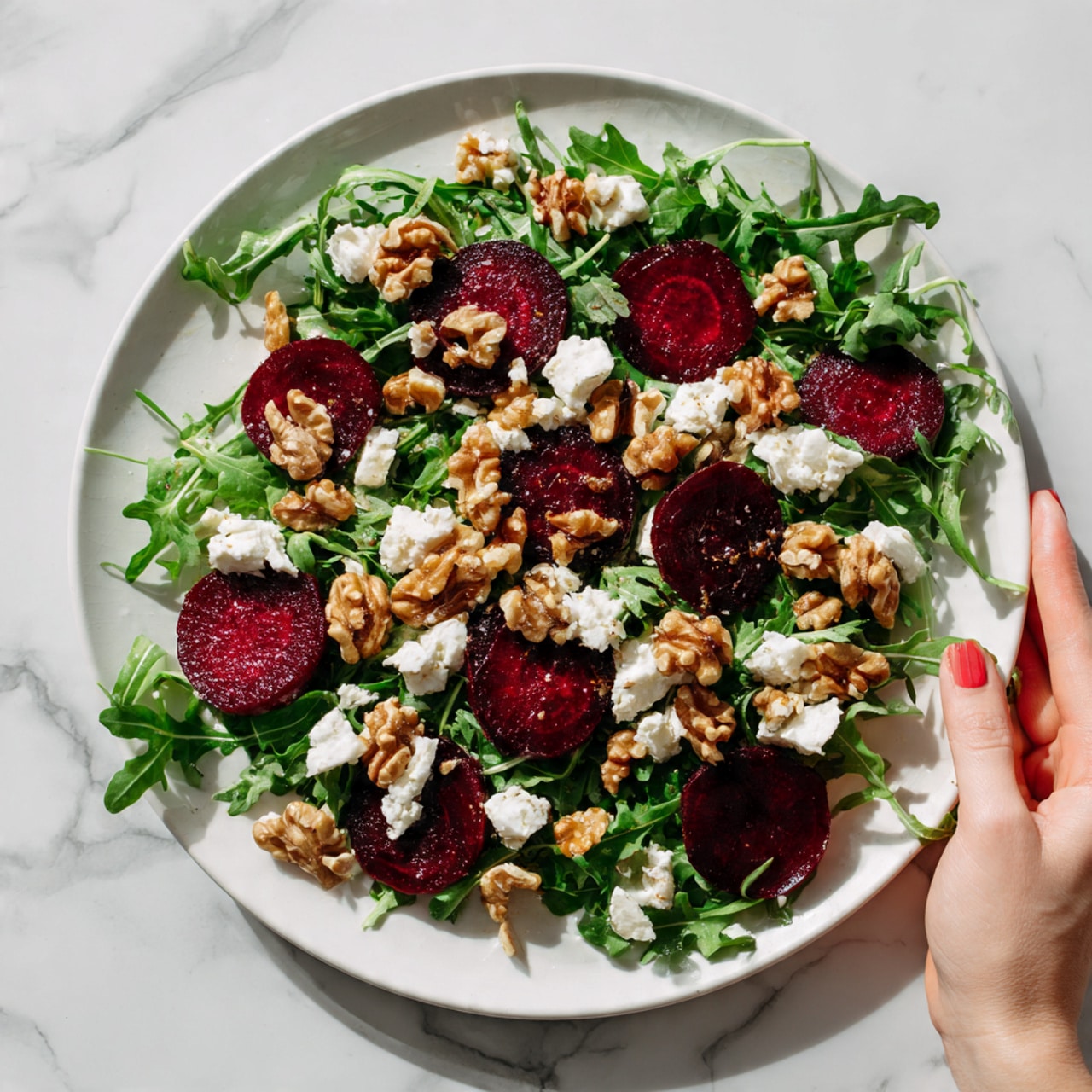 The image shows a white plate filled with a fresh salad on a white marbled surface. The salad has a base layer of green leafy arugula spread evenly across the plate. On top, there are round, deep red beet slices placed in a scattered pattern. Clusters of brown, crunchy walnuts are spread all over the salad, adding texture. Small pieces of soft white cheese are sprinkled unevenly on top, contrasting with the darker ingredients. A woman's hand is holding the edge of the plate, adding a human touch. The whole scene is softly lit, making the colors of the salad stand out vibrantly. photo taken with an iphone --ar 4:5 --v 7