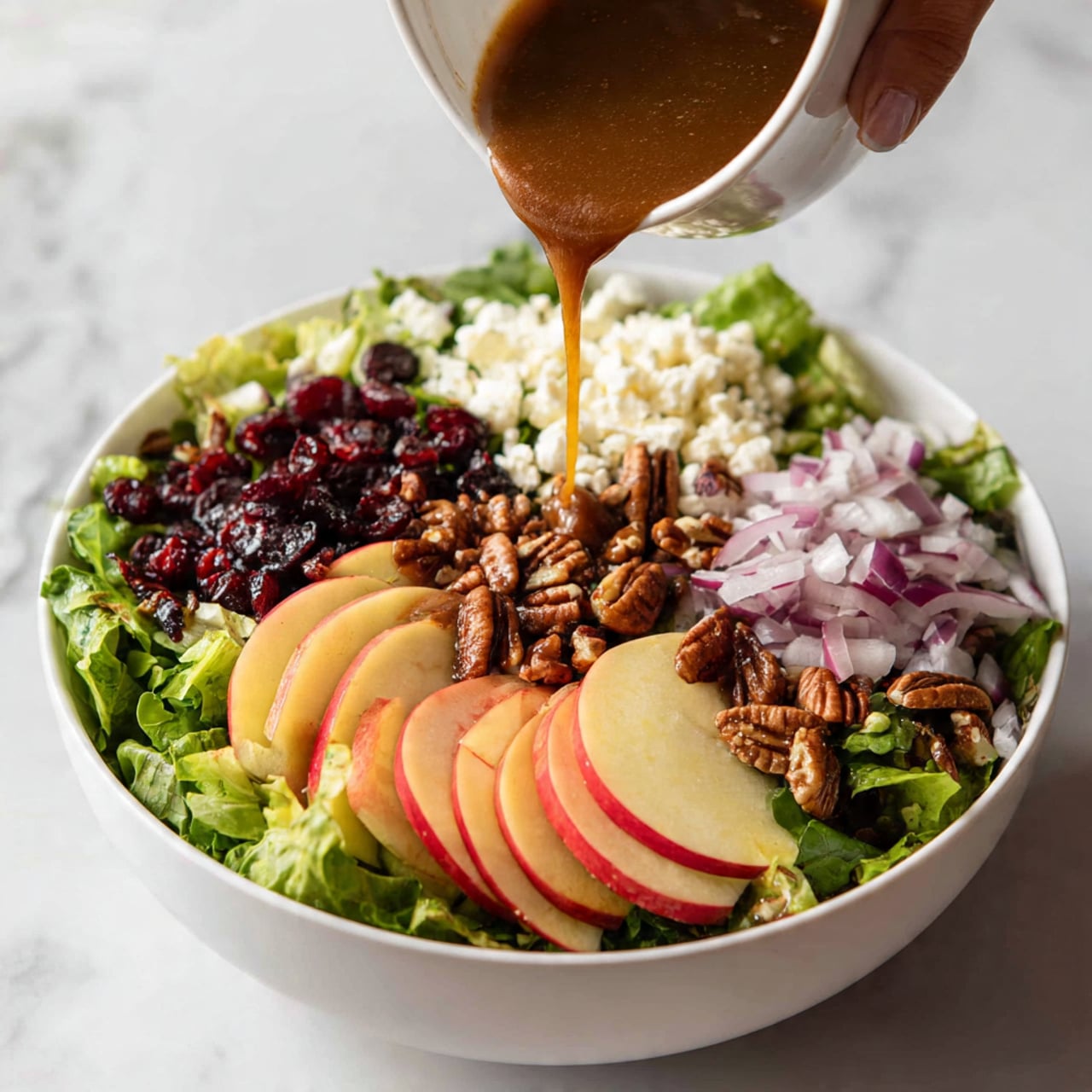 A white bowl filled with a fresh salad showing many layers: the base layer is green leafy lettuce, on top are thin slices of red and yellow apple arranged evenly. Scattered among the apples are small white cheese crumbles, dark red dried cranberries, and whole brown pecans. Red onion rings are spread across the salad, adding a vibrant splash of purple. A woman's hand is seen pouring a thick brown dressing over the top. The background is a white marbled surface. photo taken with an iphone --ar 4:5 --v 7