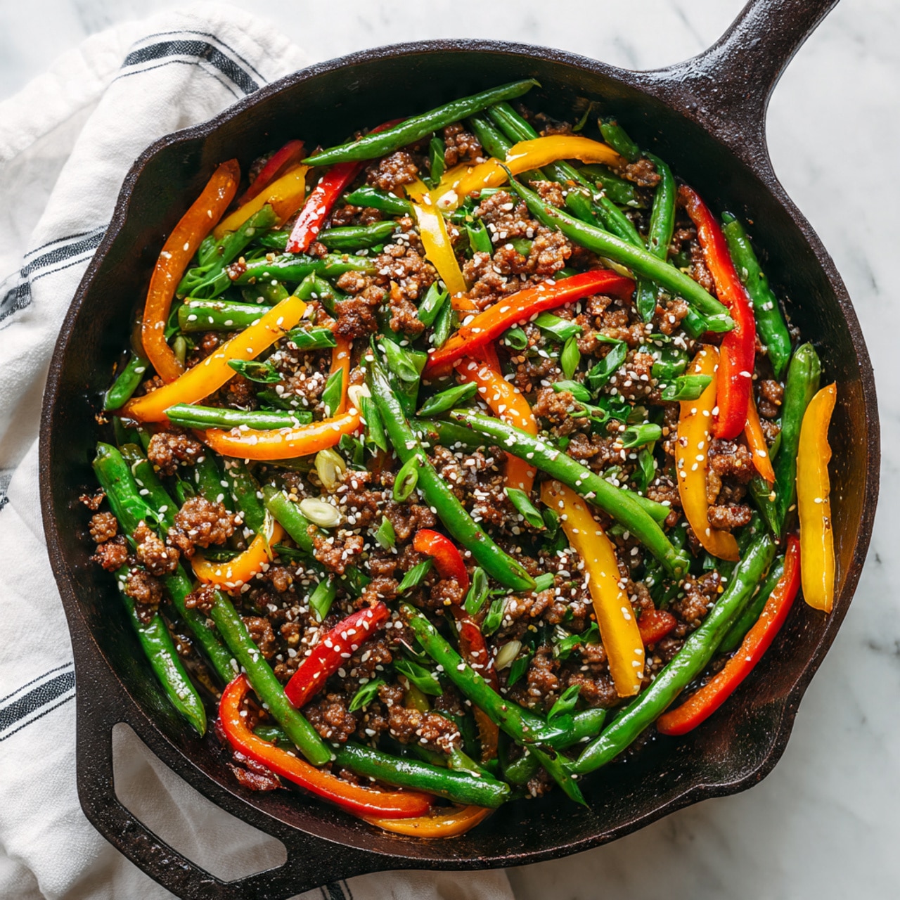 A black cast iron pan filled with a colorful stir-fry sits on a white marbled surface, layered with bright green cut green beans scattered throughout, thin bright orange carrot sticks, and yellow bell pepper strips giving a fresh pop of color. The stir fry includes small pieces of brown cooked ground meat mixed evenly, coated in a glossy sauce. The vegetables and meat are sprinkled with small white sesame seeds and chopped green onions to add texture. The pan rests on a white cloth with black simple line patterns. photo taken with an iphone --ar 4:5 --v 7