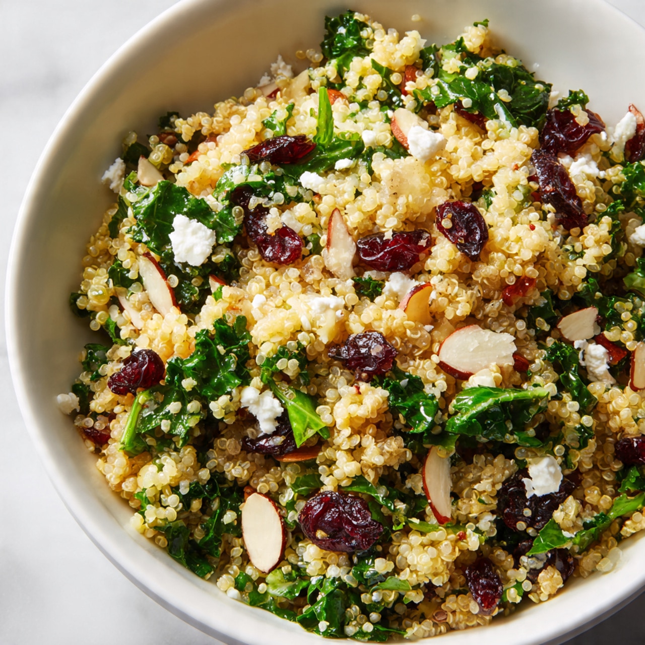 The image shows a close-up of a quinoa salad in a white bowl placed on a white marbled surface. The salad has multiple layers with small, round, pale yellow quinoa grains as the base. Scattered evenly throughout are bright green spinach leaves and chopped kale, adding a fresh leafy texture. There are also dark red dried cranberries spread over the top, along with light cream-colored almond slices that provide a crunchy look. Small bits of white feta cheese are mixed in, creating a crumbly contrast among the colors and textures. photo taken with an iphone --ar 4:5 --v 7