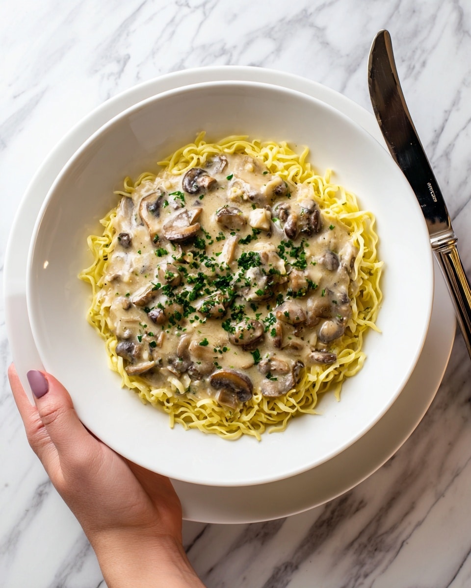 A white bowl filled with a neat layer of yellow egg noodles at the bottom, topped with a thick, creamy light beige sauce mixed with small pieces of brown mushrooms, garnished with chopped green herbs scattered on top. The bowl is placed on a matching white plate, all set on a white marbled surface. A silver knife lies to the right side of the plate, and a woman's hand is gently holding the bowl from the top right edge. photo taken with an iphone --ar 4:5 --v 7