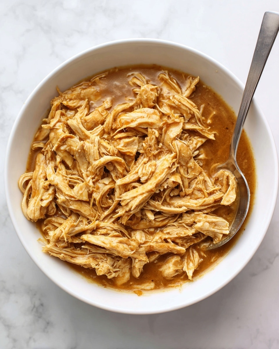 The image shows a white bowl filled with a dish of shredded cooked chicken mixed with a thick, light brown sauce. The chicken pieces are soft and moist, spread evenly in the bowl with the sauce coating every part. There is a silver spoon resting inside the bowl, placed toward the back right edge. The bowl sits on a white marbled surface, making the colors of the food stand out clearly. Photo taken with an iphone --ar 4:5 --v 7