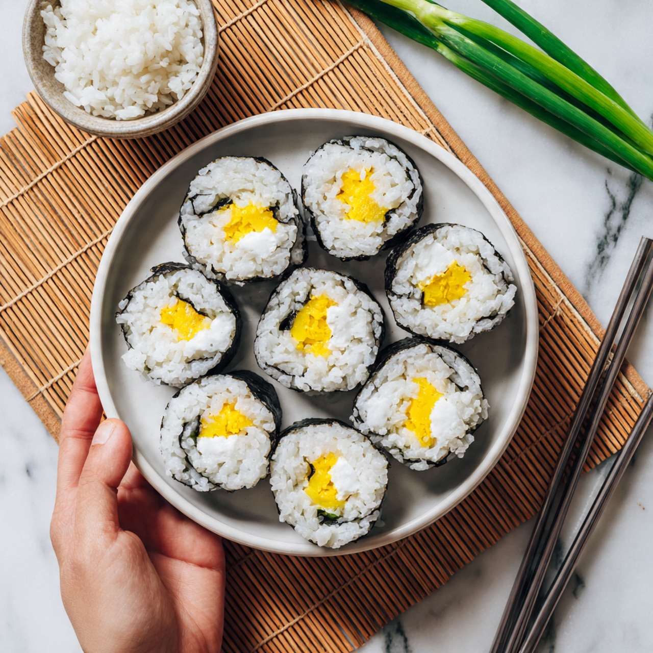 The image shows a white bowl filled with white rice topped with multiple sushi rolls arranged in a circle. Each sushi roll has a dark seaweed outer layer, followed by a thick white rice layer inside, and a bright yellow filling in the center that looks smooth and soft. The bowl is placed on a light textured bamboo mat, and a few green onion pieces and a woman's hand holding a green onion are visible at the top left. There is also a small white bowl with rice and a pair of metal chopsticks lying on the white marbled surface near the bowl. Photo taken with an iphone --ar 4:5 --v 7