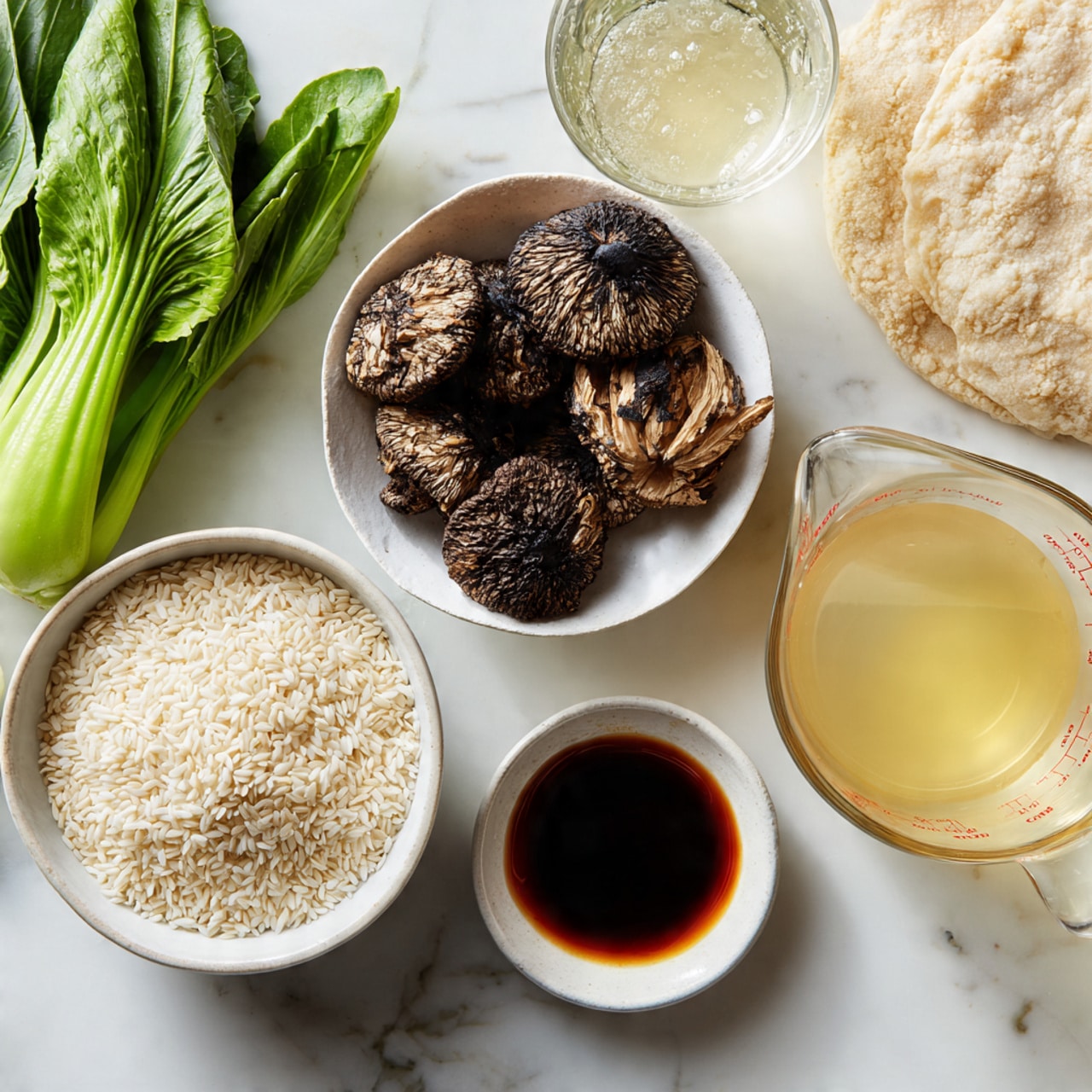 The image shows several white bowls and a glass measuring cup on a white marbled surface. One white bowl contains raw white rice grains, another holds dried brown shiitake mushrooms with a textured surface, and a smaller white bowl has a dark reddish liquid, likely soy sauce. A glass measuring cup filled with a light yellow broth or stock sits on the right side. There is also fresh green bok choy with thick stems placed on the upper left and a pale flatbread or dough in the upper right corner. The overall scene looks clean and organized, ready for cooking photo taken with an iphone --ar 4:5 --v 7