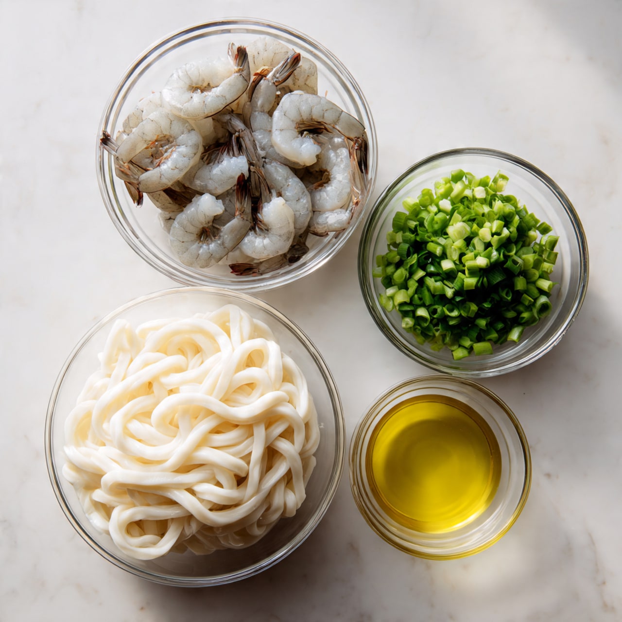 The image shows four clear glass bowls on a white marbled surface, each holding different ingredients. The largest bowl in the front contains a block of thick, white udon noodles with a soft, twisted texture. Behind it to the left is a medium bowl with several raw shrimp, pale gray with darker tails, arranged in a circle. To the right of the shrimp bowl is a small bowl filled with chopped green onions, bright green and white pieces mixed together. The smallest bowl on the far right holds a light yellow liquid, possibly oil or broth. The items are arranged neatly and the lighting highlights the clean, fresh look of each ingredient. photo taken with an iphone --ar 4:5 --v 7