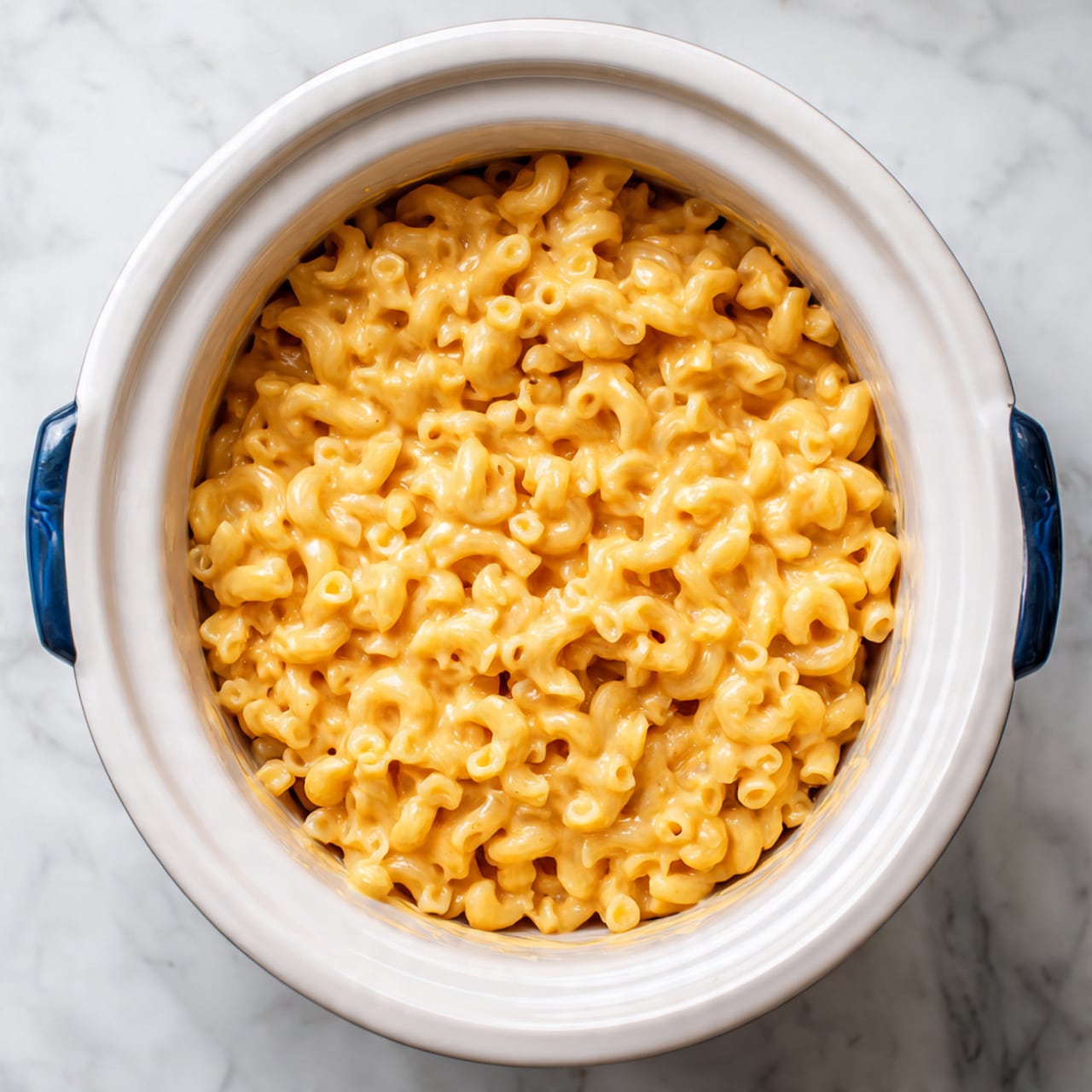 The image shows a white slow cooker filled with creamy macaroni and cheese. The macaroni noodles are elbow-shaped, soft, and covered in a smooth, light orange cheese sauce that looks thick and rich. The cheese sauce evenly coats every noodle, giving the dish a shiny, melted texture. The slow cooker has blue handles on both sides and sits on a white marbled surface. photo taken with an iphone --ar 4:5 --v 7