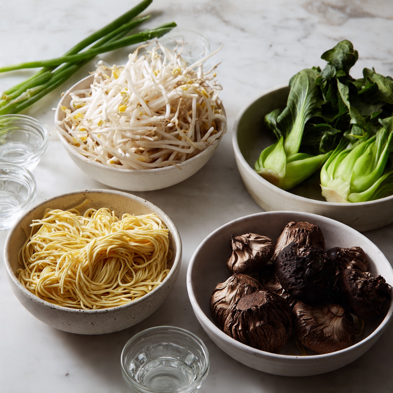 The image shows a close-up of four round white bowls arranged on a white marbled surface. The first bowl at the bottom left holds a neat pile of light yellow dry noodles with a slightly rough texture. To its right, a bowl is filled with thick, brown shiitake mushrooms with a slightly wrinkled look and dark caps. Above these, to the left, there is a bowl filled with white bean sprouts that look crisp and fresh. On the top right, a bowl contains bright green leafy vegetables and long green stems, giving a fresh and lively contrast. Three small clear glasses are visible on the left side near the bowls. A few long green onions lie across the bowls, adding a fresh green accent to the scene. The whole setup is softly lit and looks fresh and ready to cook. photo taken with an iphone --ar 4:5 --v 7