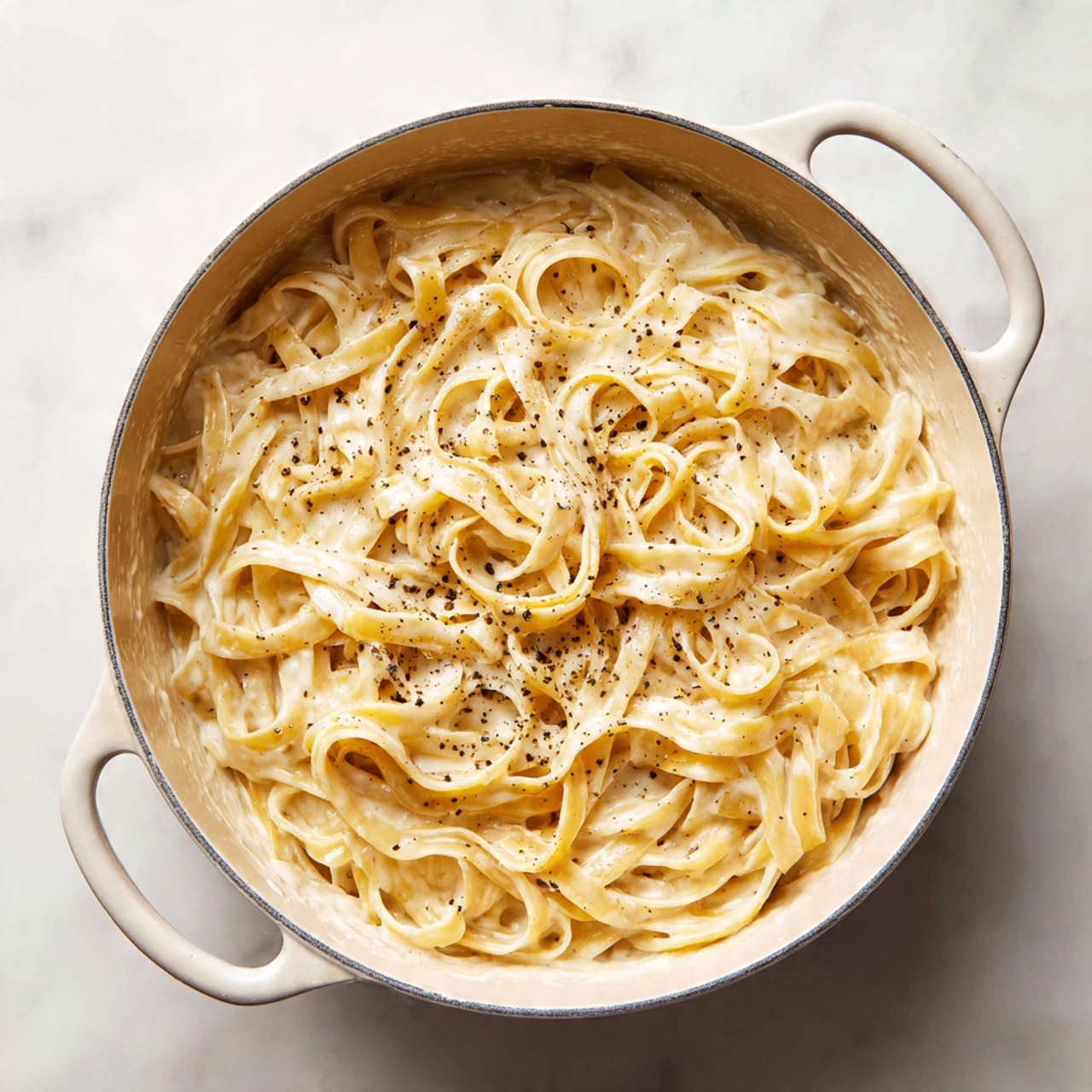 A white pot filled with creamy Alfredo fettuccine pasta shows many long, flat noodles twisted and piled unevenly inside. The thick sauce is light beige with a smooth texture, lightly speckled with black pepper. The pot sits on a white marbled surface, and the edges of the pot are softly rounded with a visible handle on one side. photo taken with an iphone --ar 4:5 --v 7