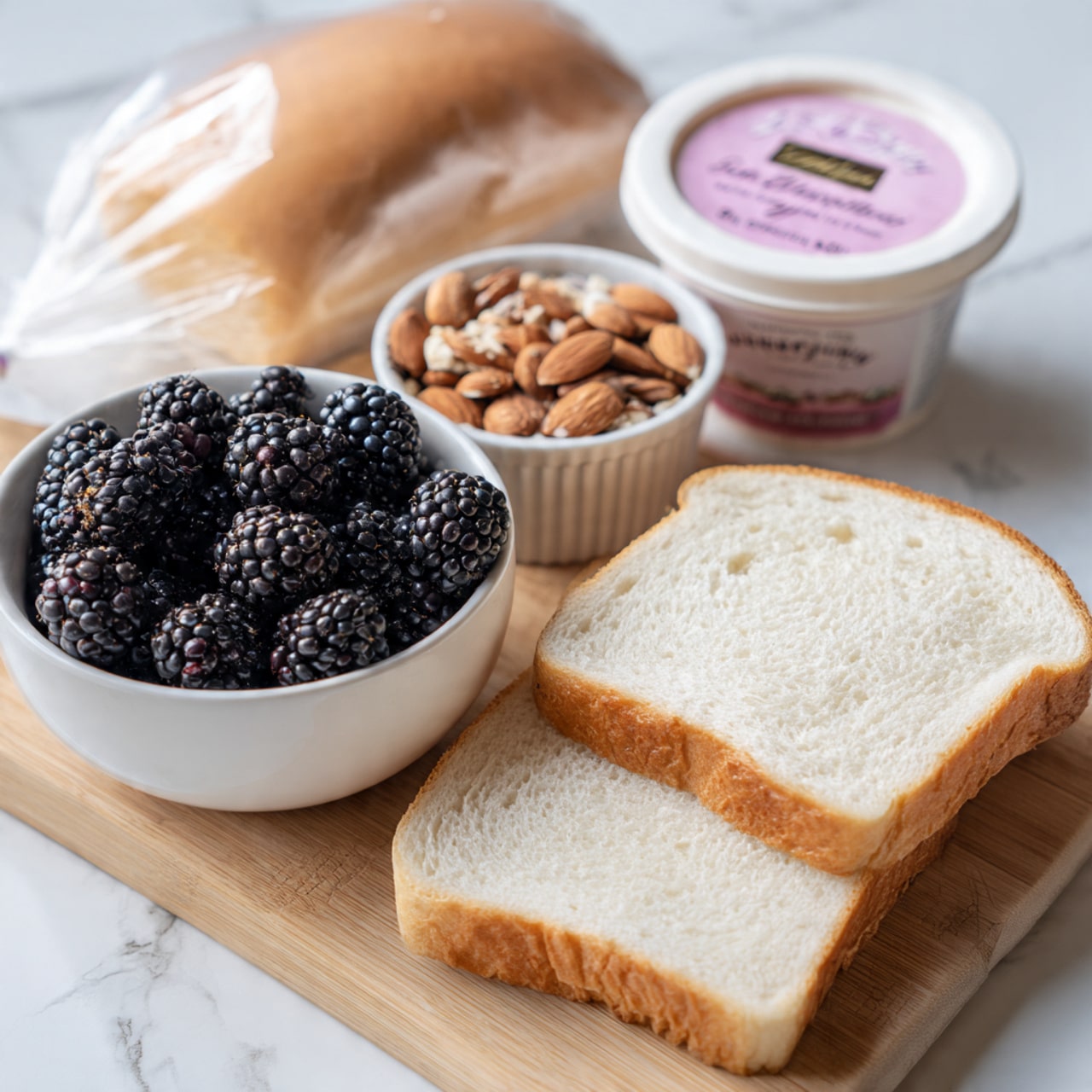 The image shows two slices of white bread lying flat on a wooden board. Next to the bread, there is a white bowl filled with dark blackberries that look fresh and plump. Beside the bowl is a small white ramekin filled with chopped almonds, showing a mix of light brown and tan colors with some rough texture. Behind these items, there is a small container of pink cream cheese with a berry flavor label, and in the background, a loaf of white sandwich bread in a clear plastic bag is partially visible. The scene is set on a white marbled surface. photo taken with an iphone --ar 4:5 --v 7