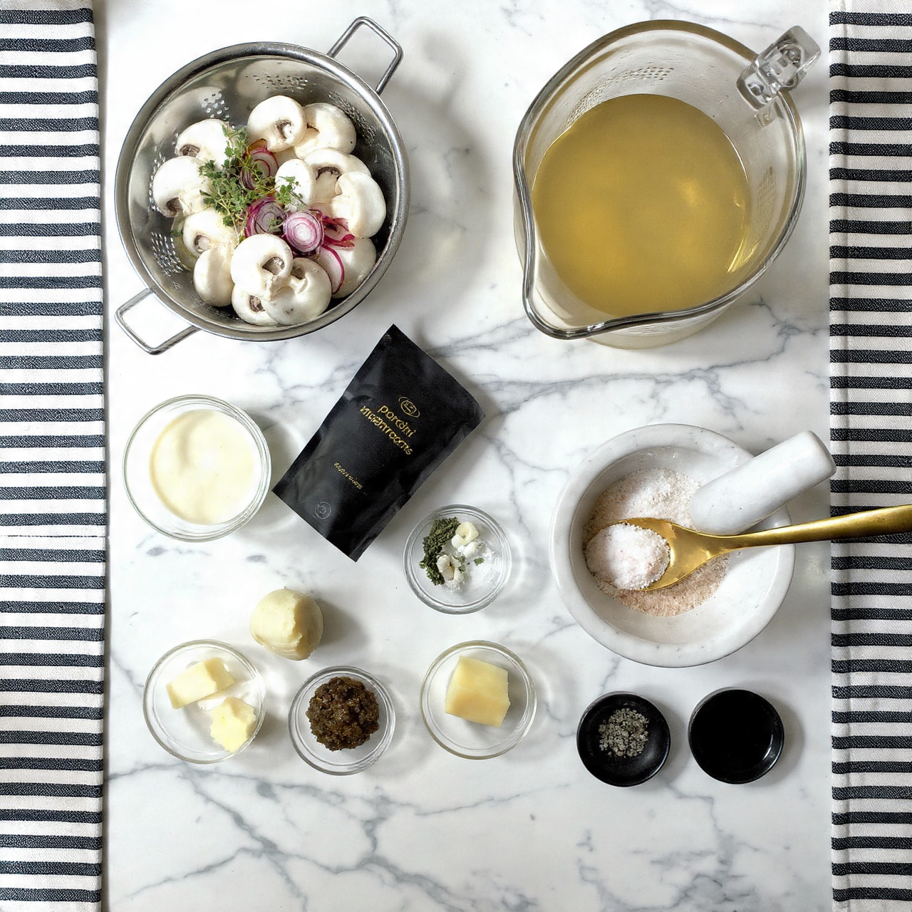 A top view shows a white marbled surface with ingredients neatly arranged for cooking. On the left side, a silver colander holds fresh white mushrooms with reddish shallots and sprigs of green thyme on top. Below it is a clear jar filled with a white creamy liquid. To the right, a clear glass gravy boat contains light brown broth. In front of it is a dark package labeled “porcini mushrooms.” Small glass bowls hold different ingredients: three garlic cloves, a pat of yellow butter, a light brown paste, a reddish brown liquid, and dark soy sauce. A white mortar contains pink salt with a golden spoon, while two small black bowls hold dried herbs and pepper. A black and white striped cloth is on both the left and right edges of the frame. photo taken with an iphone --ar 4:5 --v 7