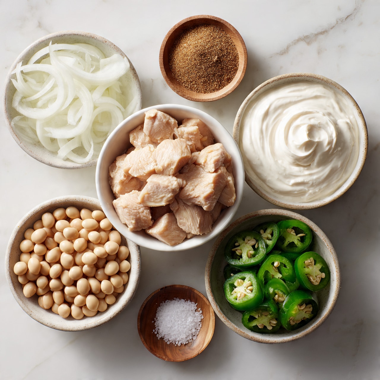 The image shows six white bowls arranged closely on a white marbled surface. In the center bowl, there are light pink cooked chicken chunks with a smooth, slightly moist texture. To the top left, a bowl filled with chopped white onions features translucent slicing with layered rings. To the bottom left, a bowl contains light beige soybeans that are shiny and smooth. On the bottom right, a bowl is filled with sliced fresh green jalapeño peppers, showing their rings and seeds inside. At the top right, a bowl of thick white cream has a swirled, soft texture. In the small bowl near the center top, there is a brown dry spice powder, and in another tiny wooden bowl near the center, there is coarse white salt. All are captured in soft natural light. Photo taken with an iphone --ar 4:5 --v 7