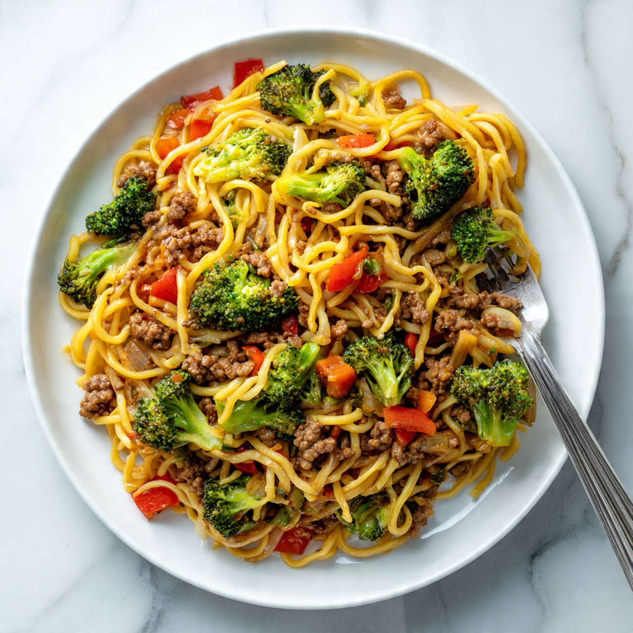 A white plate filled with cooked noodles mixed evenly with small pieces of broccoli, diced red bell peppers, and ground meat, all layered together creating a colorful mix of yellow noodles, green broccoli, and red peppers. The texture looks soft and slightly oily, with a silver fork resting inside the noodles on the right side of the plate. The background is a white marbled surface, and the lighting highlights the glossy shine on the food, showing the fresh vegetables and tender meat. Photo taken with an iphone --ar 4:5 --v 7