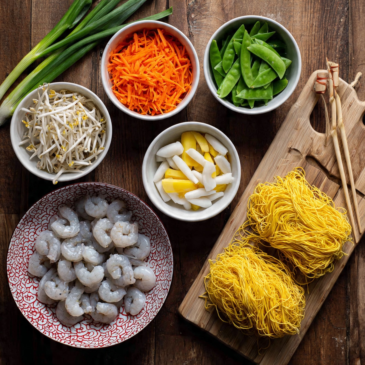 The image shows several bowls and a wooden board on a dark wooden surface, with ingredients arranged for cooking. A white bowl with a red geometric pattern holds raw peeled shrimp, pale gray with tails on the outer side. Next to it on the right are wooden chopsticks. On the wooden board lies a neat bundle of yellow uncooked noodles with a smooth texture. Around these are five white bowls with different vegetables: bright orange shredded carrots in one bowl, white round slices in another, light yellow rectangular slices in a third, fresh green snap peas in a fourth, and pale white bean sprouts in the last bowl. Three green onions rest on the surface at the top left. The entire setup is on a white marbled texture surface, and the image feels organized and ready for cooking photo taken with an iphone --ar 4:5 --v 7