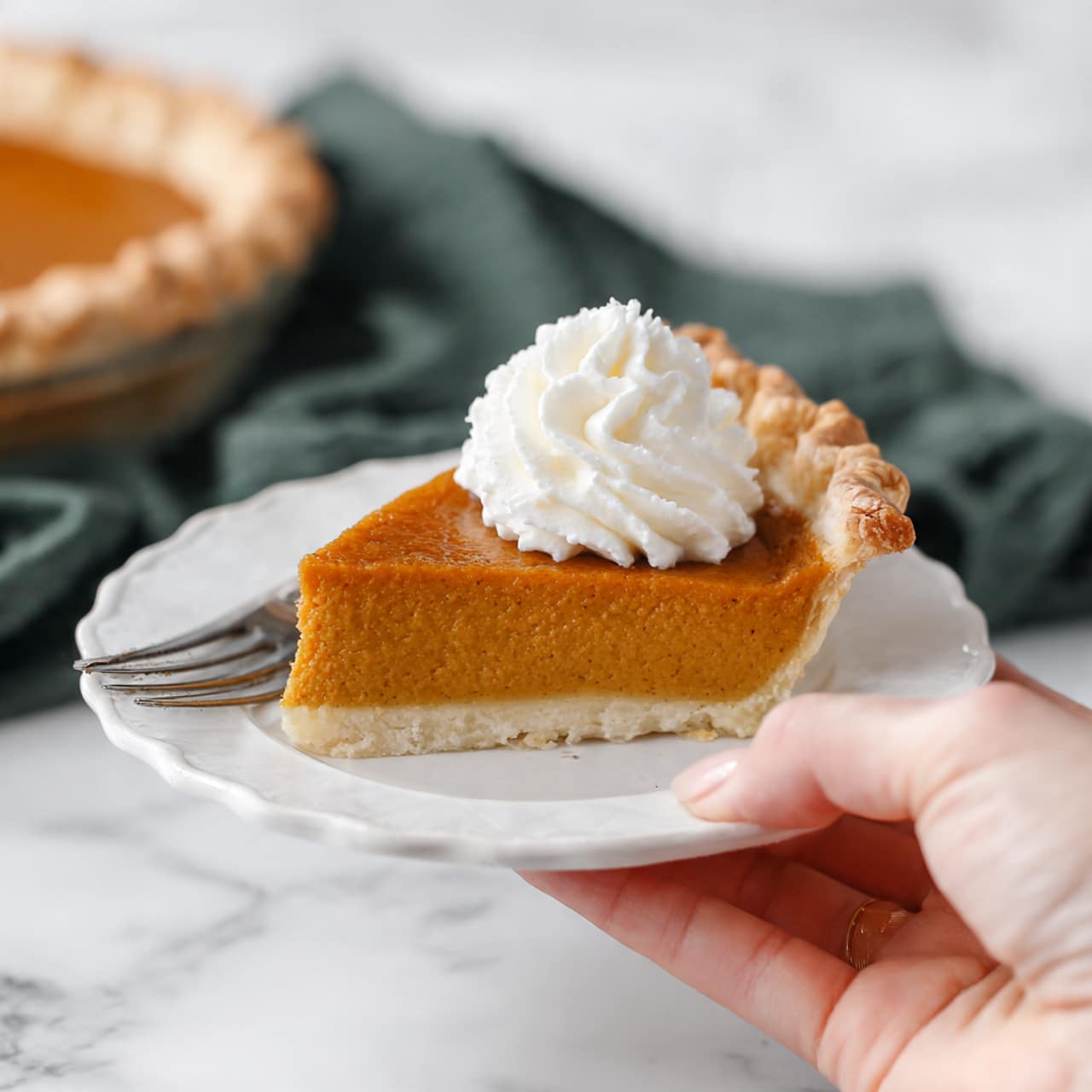 A slice of pumpkin pie with a smooth orange filling and a golden-brown flaky crust sits on a small white plate with a scalloped edge. On top of the pie slice is a swirl of white whipped cream. The white marbled surface beneath the plate adds a clean background, and a silver fork rests beside the plate. A woman's hand holds the edge of the plate gently. In the background, part of the pie and a dark green cloth napkin are visible. Photo taken with an iphone --1:1