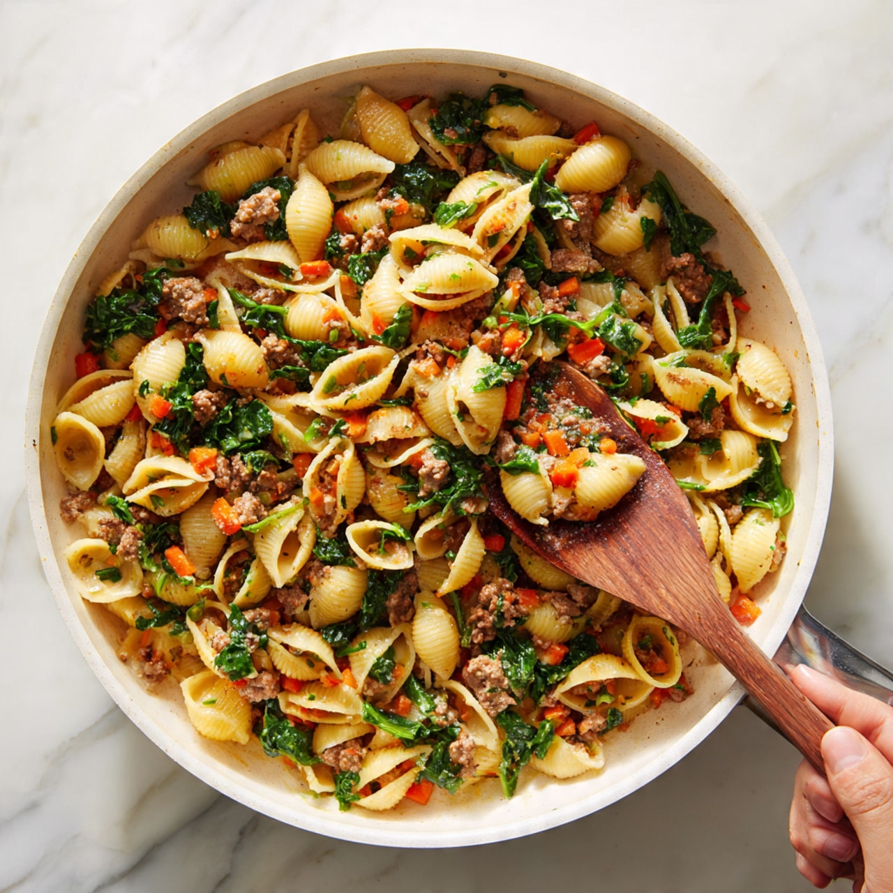 A white pan filled with a cooked pasta dish made of shell-shaped pasta mixed with small pieces of cooked meat and green leafy vegetables. There are diced orange carrot pieces spread throughout, adding a pop of color. A woman's hand holds a wooden spatula scooping some of the pasta, showing the dish's mixed textures and layers. The background is a white marbled surface. photo taken with an iphone --ar 4:5 --v 7