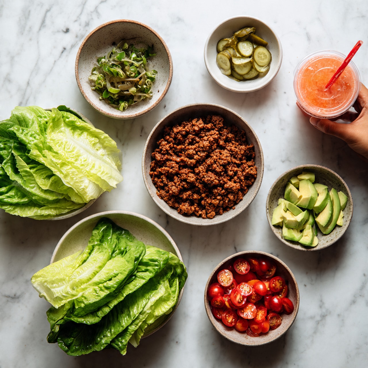 The image shows a white marbled surface with six bowls arranged in a loose circle. Two bowls contain fresh, bright green lettuce leaves with some darker green edges, positioned at the bottom left and right. Above them, a bowl filled with cooked ground meat, brown and crumbly, is placed on the top left. To the right of that, a small bowl with sliced green pickles is near the top center. Below it, a bowl with fresh, sliced avocado pieces in light green is visible near the center. A small bowl with halved, bright red cherry tomatoes is beside the avocado on the right side. Next to the tomatoes, there is a white bowl with a small amount of pinkish-orange dressing. A woman’s hand holding a red drink cup with a straw is visible in the top right corner. Photo taken with an iphone --ar 4:5 --v 7