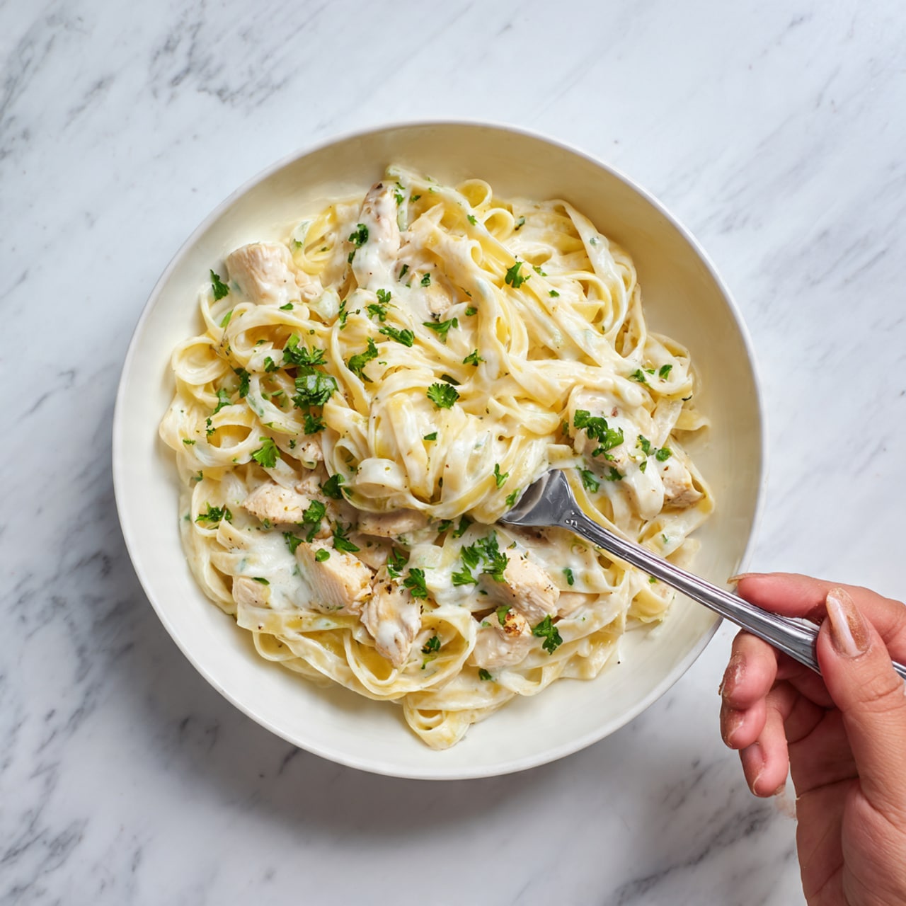 The image shows a white bowl filled with creamy fettuccine pasta. The pasta is thick and coated evenly with a smooth white sauce that looks rich and shiny. Small pieces of light-colored cooked chicken are mixed into the pasta, with a few green parsley leaves on top for color. A silver fork is twirling some pasta in the center, and a woman's hand holds the fork. The bowl is placed on a white marbled surface. photo taken with an iphone --ar 4:5 --v 7