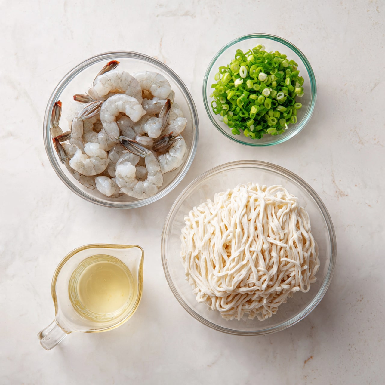 The image shows four clear glass bowls on a white marbled surface, each with different ingredients. The largest bowl at the bottom right holds a block of thick, white noodles with a soft, slightly shiny texture. To the left, a medium bowl contains several raw, peeled shrimp, light gray with darker tails, resting closely together. Above the shrimp, a smaller bowl has fresh chopped green onions, bright green and finely sliced. Next to it on the right, a small measuring cup holds a pale yellow liquid, likely oil or broth. The setup is clean and organized, ready for cooking. photo taken with an iphone --ar 4:5 --v 7