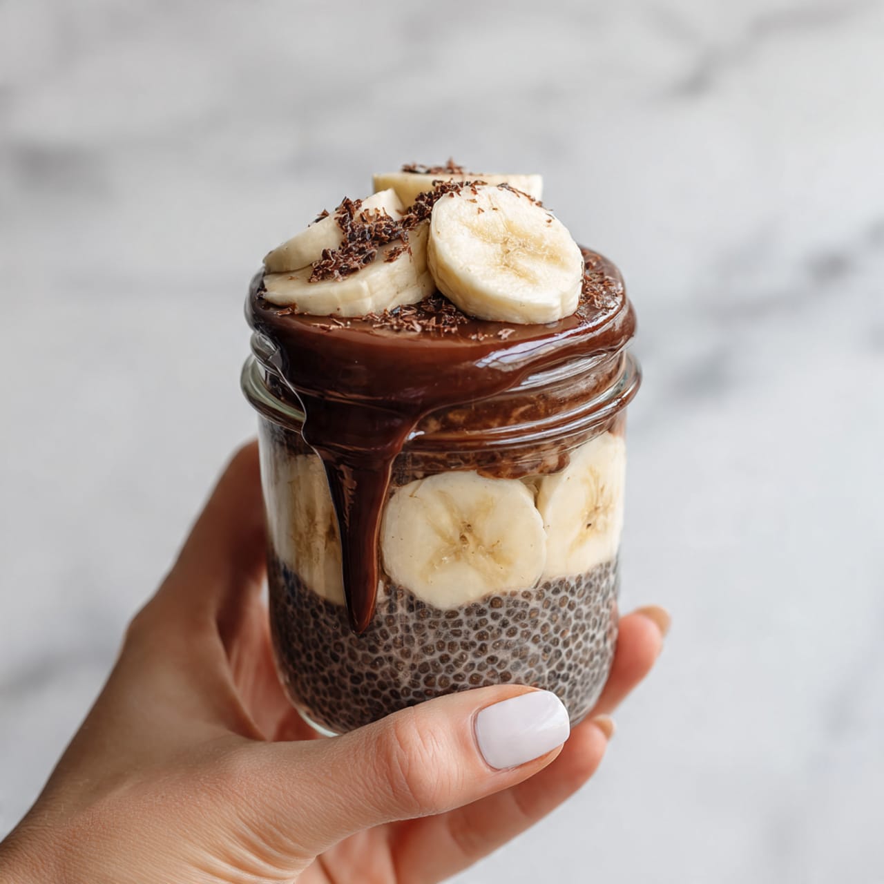 A clear glass jar held by a woman's hand contains a layered dessert with four main layers. The bottom layer is dark brown and textured, likely chocolate chia pudding. Above this is a layer of sliced banana pieces in a pale cream color. Another layer of the same dark brown chia pudding follows the bananas. The top layer is a light creamy beige, topped with shiny, thick chocolate sauce spilling slightly down the side of the jar. The jar is set against a white marbled surface. photo taken with an iphone --ar 4:5 --v 7