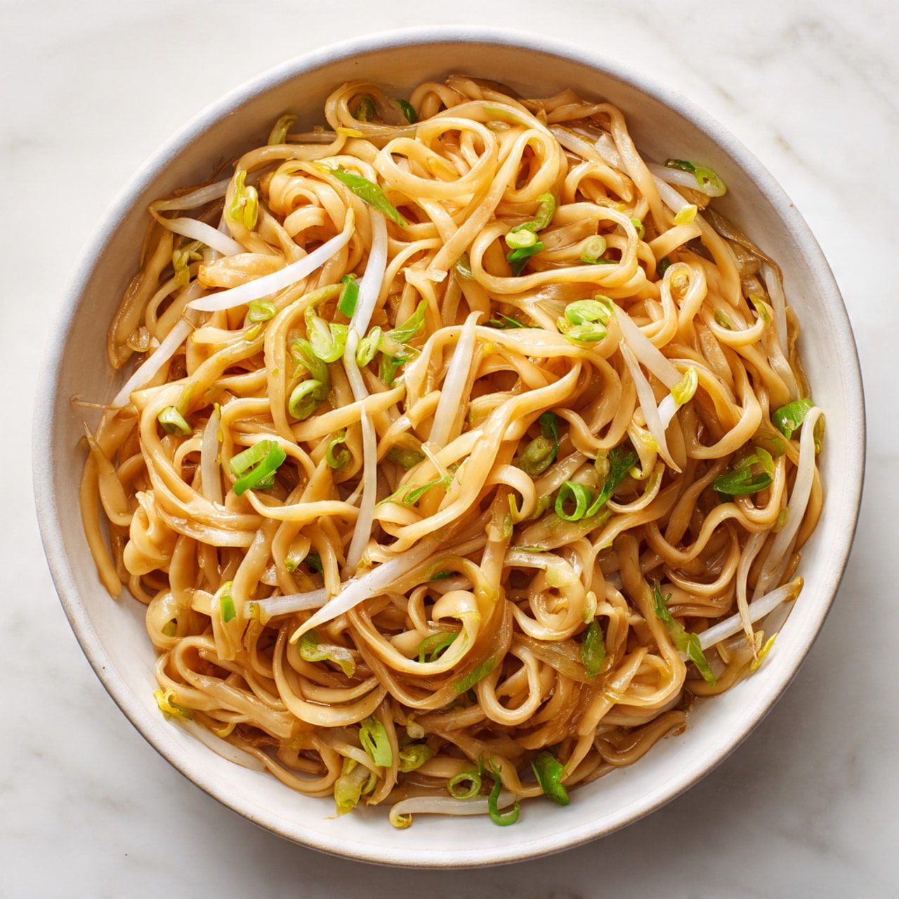 A close-up of a white bowl filled with a nest of thick, golden-brown noodles mixed with thin, white bean sprouts and small pieces of green onion scattered throughout. The noodles are glossy and look soft, twisted together in the center, with some bean sprouts and green onions peeking through. The bowl sits on a white marbled surface, giving a clean and fresh background. photo taken with an iphone --ar 4:5 --v 7