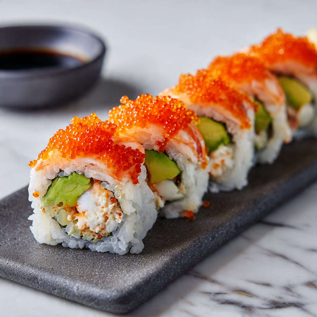 A row of four sushi rolls with white rice on the outside, each roll topped with bright orange fish roe. Inside the rolls, there is visible green avocado and white crab meat. Two cooked shrimp tails stand upright at the back, adding height and texture to the presentation. The sushi is placed on a rectangular dark gray slate plate on a white marbled background. In the background, a small black bowl with soy sauce is partly visible. photo taken with an iphone --ar 4:5 --v 7