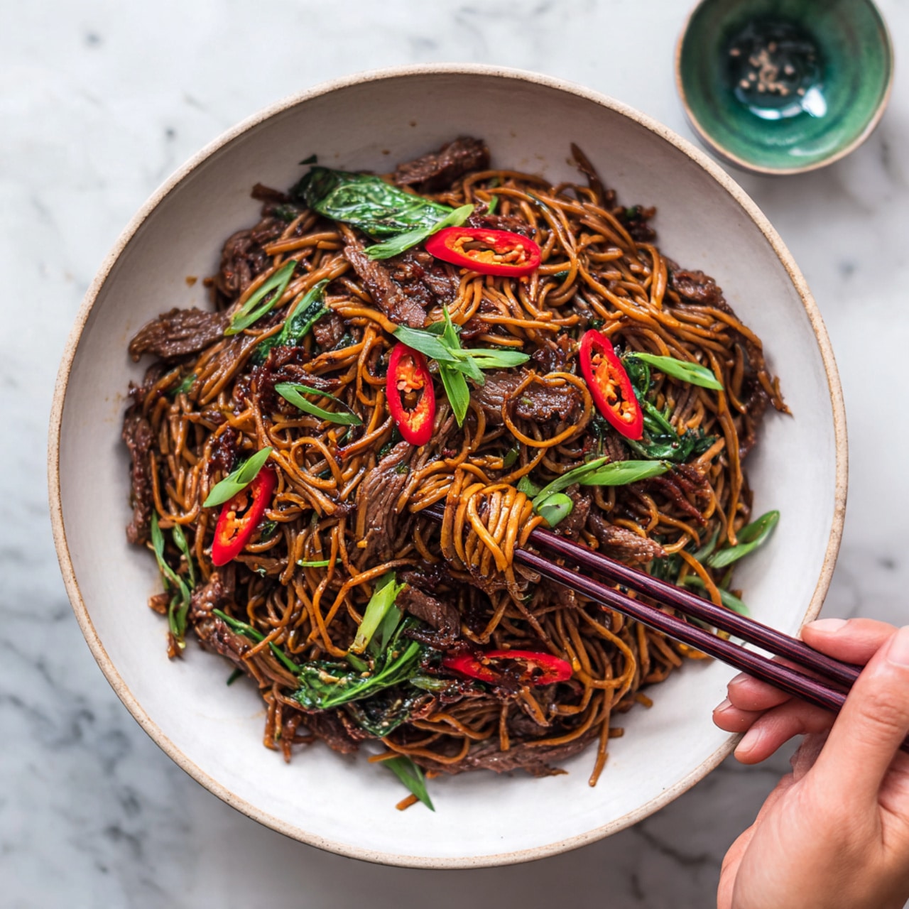 A round white bowl filled with dark brown stir-fried noodles, mixed with long red chili slices and green leaves scattered on top. Thin dark brown strips of meat are spread evenly throughout the noodles. A woman's hand holding dark brown chopsticks is picking up some noodles from the bowl. The bowl sits on a white marbled surface with a blurred small green bowl in the background. photo taken with an iphone --ar 4:5 --v 7