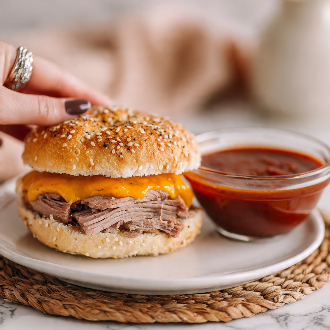 The image shows a sandwich on a white sesame seed bun with layers of thinly sliced roast beef and melted orange cheddar cheese, placed on a white plate on a white marbled surface. In the foreground, there is a clear glass bowl filled with thick red sauce, sitting on a woven mat. A woman's hand with a shiny ring holds the sandwich from the top left. The scene is lit softly, focusing on the sandwich and sauce, giving a fresh and inviting look. photo taken with an iphone --ar 4:5 --v 7