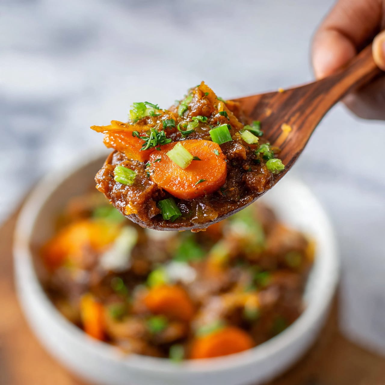 A close-up view of a wooden spoon held by a woman's hand, gently lifting a serving of a dish with three visible layers: a top layer of golden-brown caramelized meat with a glossy texture, a middle layer of soft, bright orange cooked carrot slice, and a few small pieces of light green chopped scallion scattered on top. In the blurred background, there is a white bowl filled with more of the same dish. The surface beneath is a white marbled texture. Photo taken with an iphone --ar 4:5 --v 7