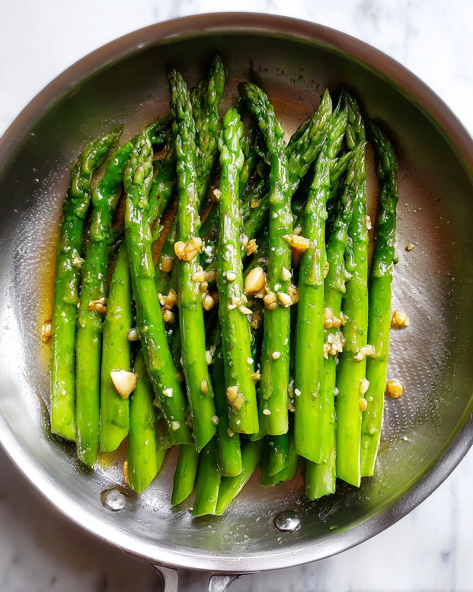 A silver pan filled with a neat row of bright green asparagus stalks, cooked to a tender texture. The asparagus is sprinkled with small bits of light golden garlic pieces, and a light sheen of oil adds a glossy finish to the stalks. The pan sits on a white marbled surface, enhancing the fresh vibrant green of the asparagus. photo taken with an iphone --ar 4:5 --v 7