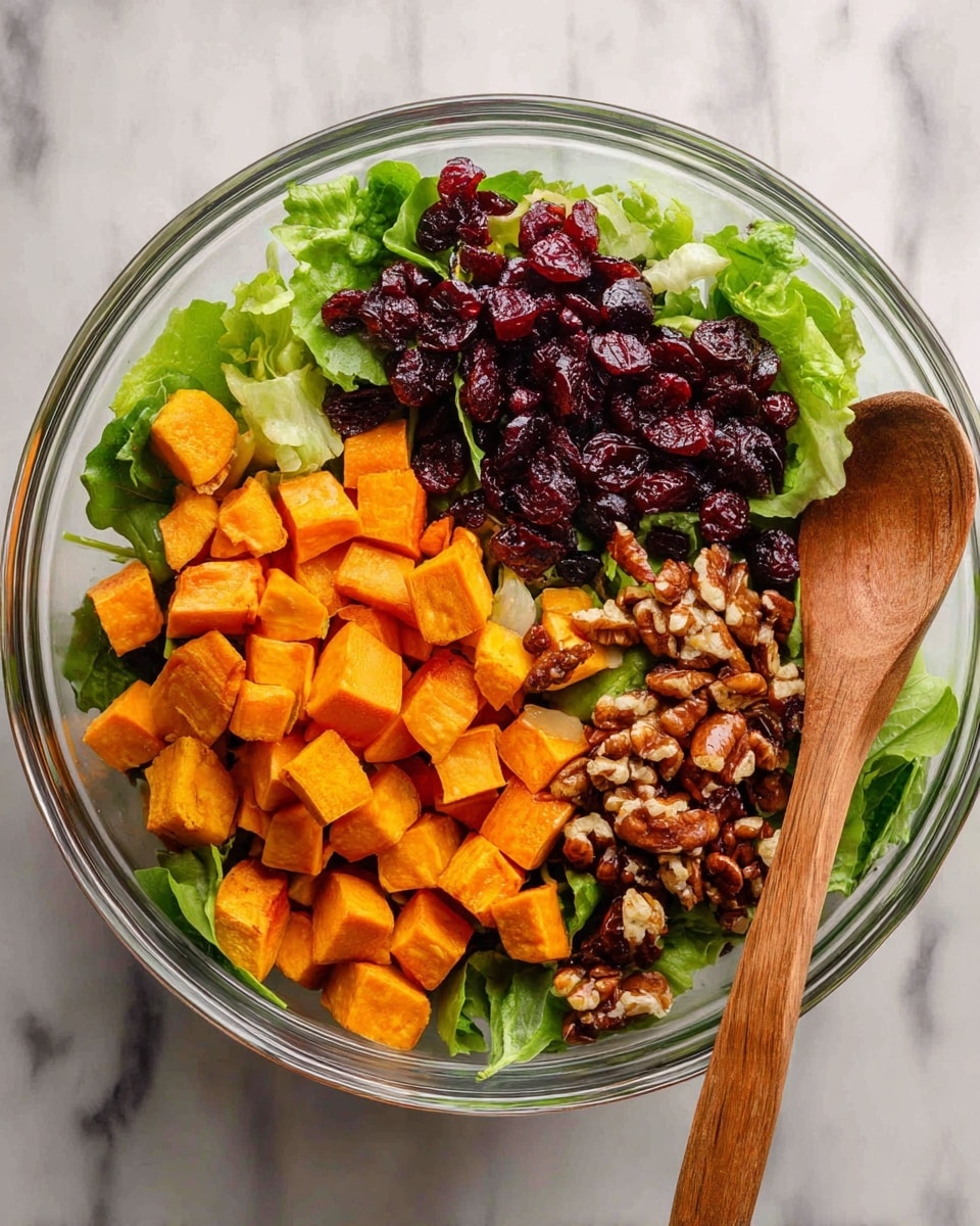 The image shows a clear glass bowl filled with a colorful salad on a white marbled surface. The bottom layer is fresh green lettuce, topped with bright orange cubed sweet potatoes piled on one side. Next to the sweet potatoes, there are dark brown roasted nuts and some dark red dried cranberries. A wooden spoon rests inside the bowl, leaning on the right side. The colors are vibrant and the textures vary from crunchy to soft. Photo taken with an iphone --ar 4:5 --v 7