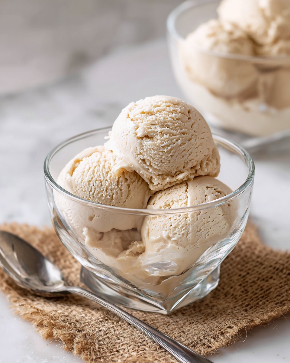 The image shows a clear glass bowl filled with three scoops of light tan ice cream with a creamy and slightly textured surface. The bowl sits on a square piece of burlap cloth, placed on a white marbled surface. Behind it, there is a second similar bowl, partly blurred, also filled with ice cream. A silver spoon rests beside the first bowl on the burlap. The lighting is soft and natural, highlighting the creamy texture of the ice cream. Photo taken with an iphone --ar 4:5 --v 7