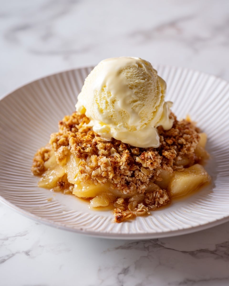 A scoop of pale yellow vanilla ice cream sits on top of a single serving of apple crumble. The crumble layer is golden brown and textured with pieces of oats and nuts, covering a thick layer of soft, cooked apple slices that look juicy and tender. This dessert is served on a white plate with a subtle raised pattern around its edge, set against a white marbled surface. The photo taken with an iphone --ar 4:5 --v 7