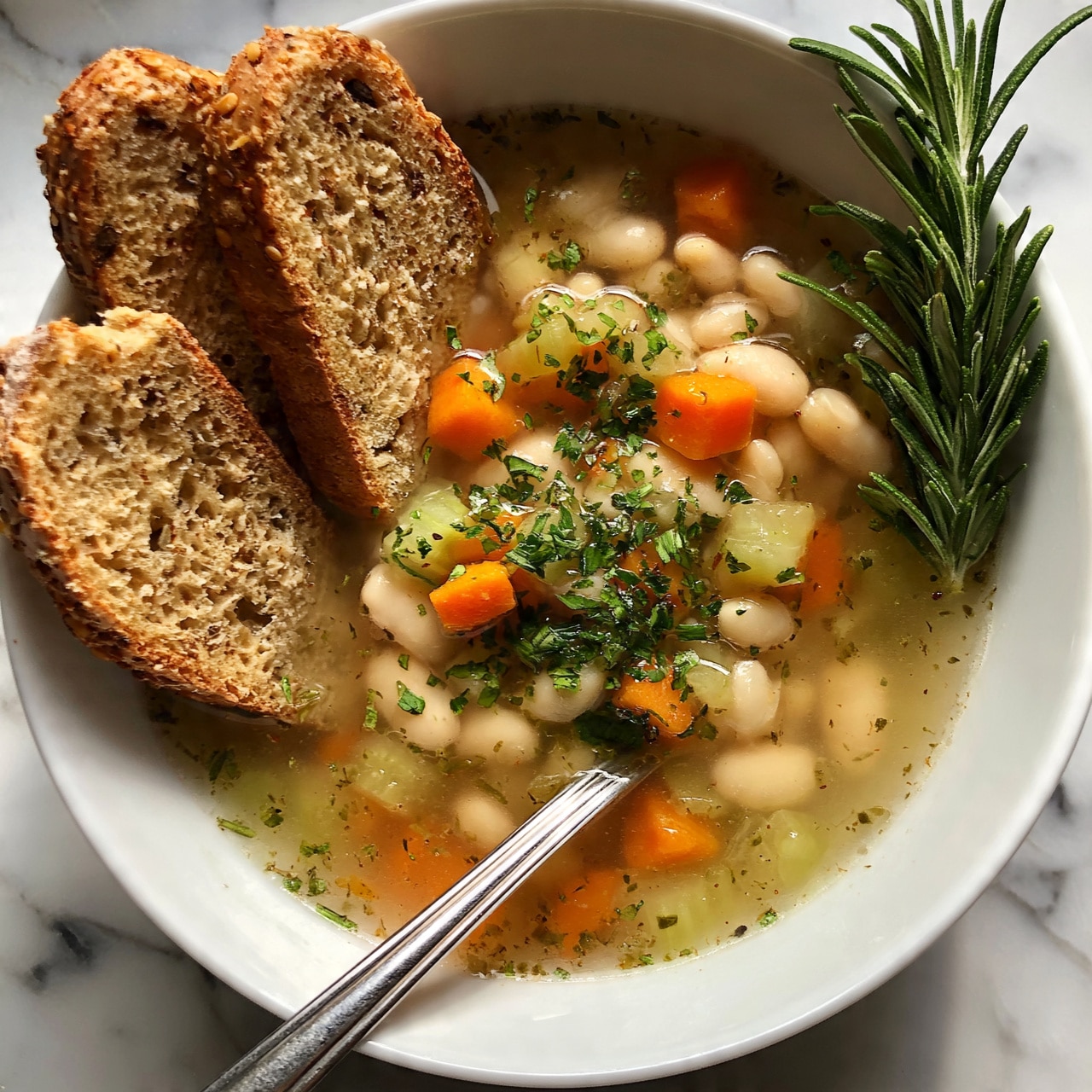 square imag A white bowl filled with clear broth soup that has white beans, small orange carrot cubes, and light green celery pieces mixed throughout, topped with chopped green herbs and a sprig of fresh rosemary resting on the right side. Three slices of toasted whole grain bread with visible seeds stand upright on the left side of the bowl. A silver spoon is angled inside the soup near the bottom center. The bowl is set on a white marbled surface. photo taken with an iphone --ar 4:5 --v 7