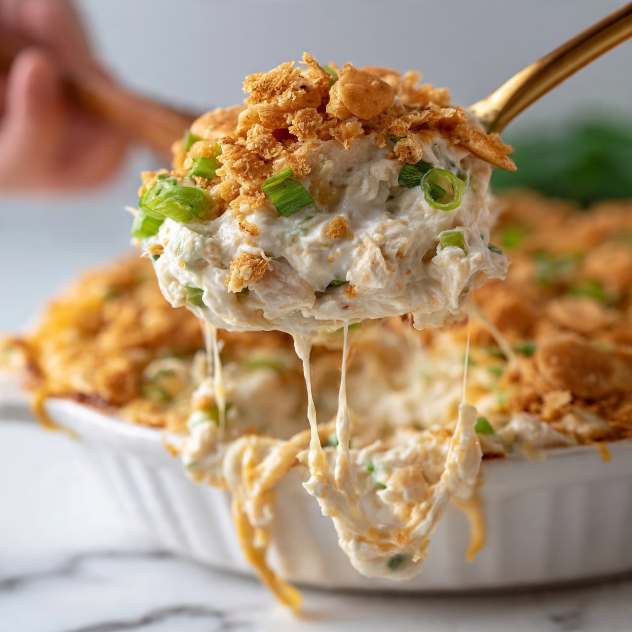 square image A close-up view shows a creamy white mixture loaded with small chunks of chicken and pieces of green onion, topped with golden crunchy cracker crumbs. The mixture looks thick and cheesy, with some gooey cheese strands stretching from the white baking dish below. A woman's hand holds a wooden spoon lifting the mixture, emphasizing the gooey texture. The dish sits on a white marbled surface in the background, with a soft, blurred green hint behind it. photo taken with an iphone --ar 4:5 --v 7square image
