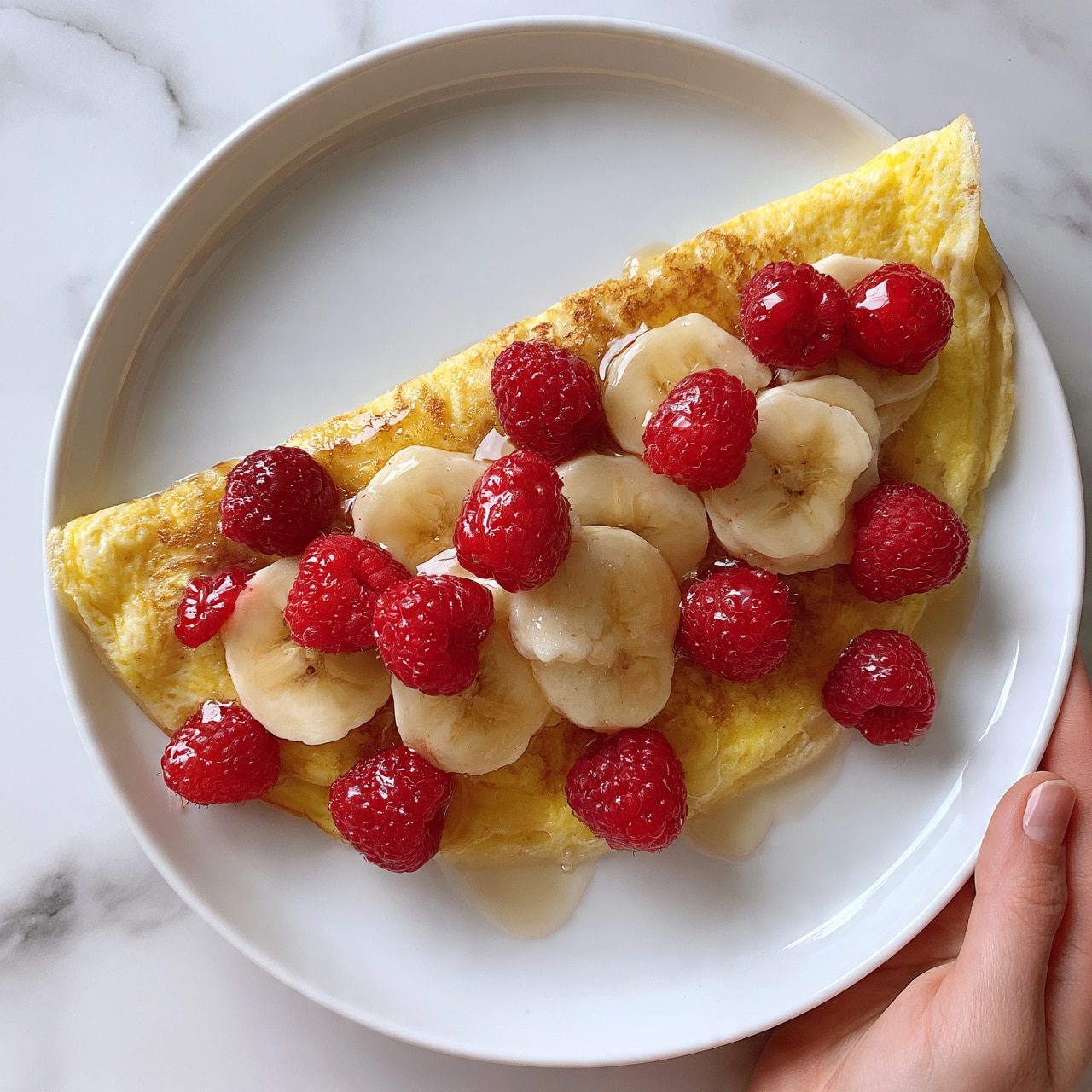square image A white plate holds a folded yellow omelet as the base layer, topped with a mix of fresh banana slices and red raspberries scattered evenly across the top and around the edges. A light drizzle of clear syrup glistens over the fruit and omelet, adding shine and texture. The background features a white marbled surface, and a woman's hand is seen gently holding the edge of the plate. Photo taken with an iphone --ar 4:5 --v 7