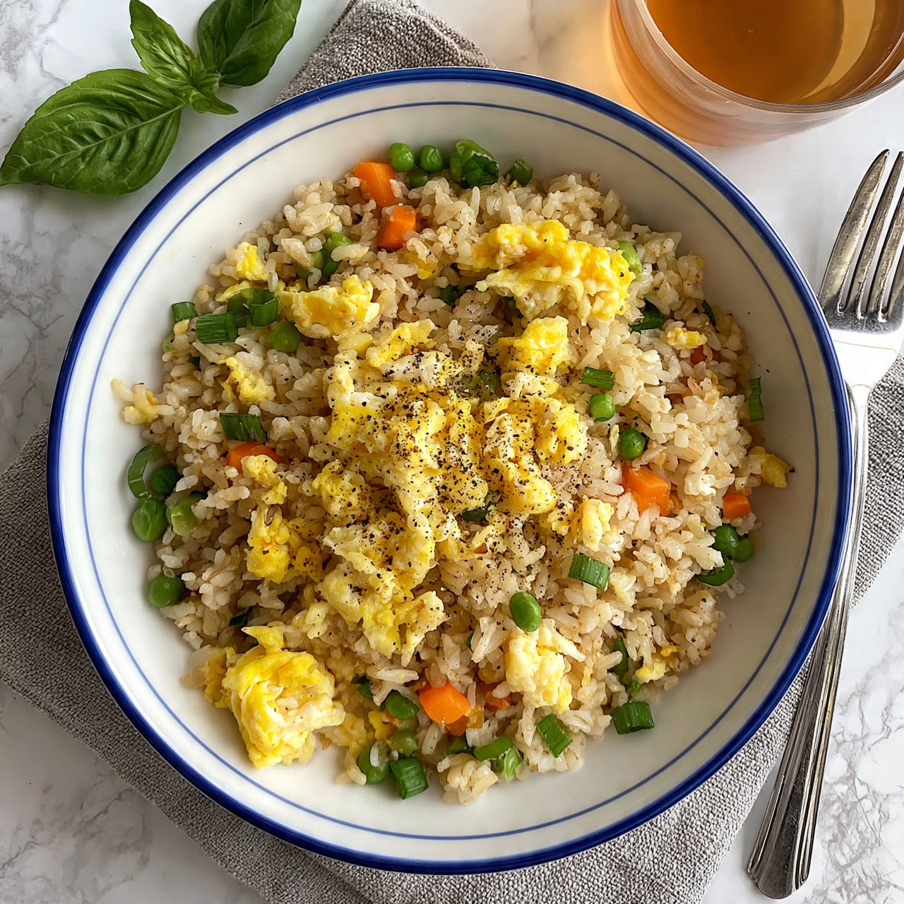 square image A white bowl with a blue rim is filled with fried rice that has three main layers visible: light brown cooked rice mixed with small bright green peas, orange diced carrots, and finely chopped green onions; scattered fluffy yellow scrambled eggs; and a sprinkling of black pepper on top. The bowl sits on a coarse gray cloth on a white marbled surface. Next to the bowl, there is a silver fork and a glass of light brown tea, with two fresh green basil leaves placed nearby. The photo taken with an iphone --ar 4:5 --v 7square image