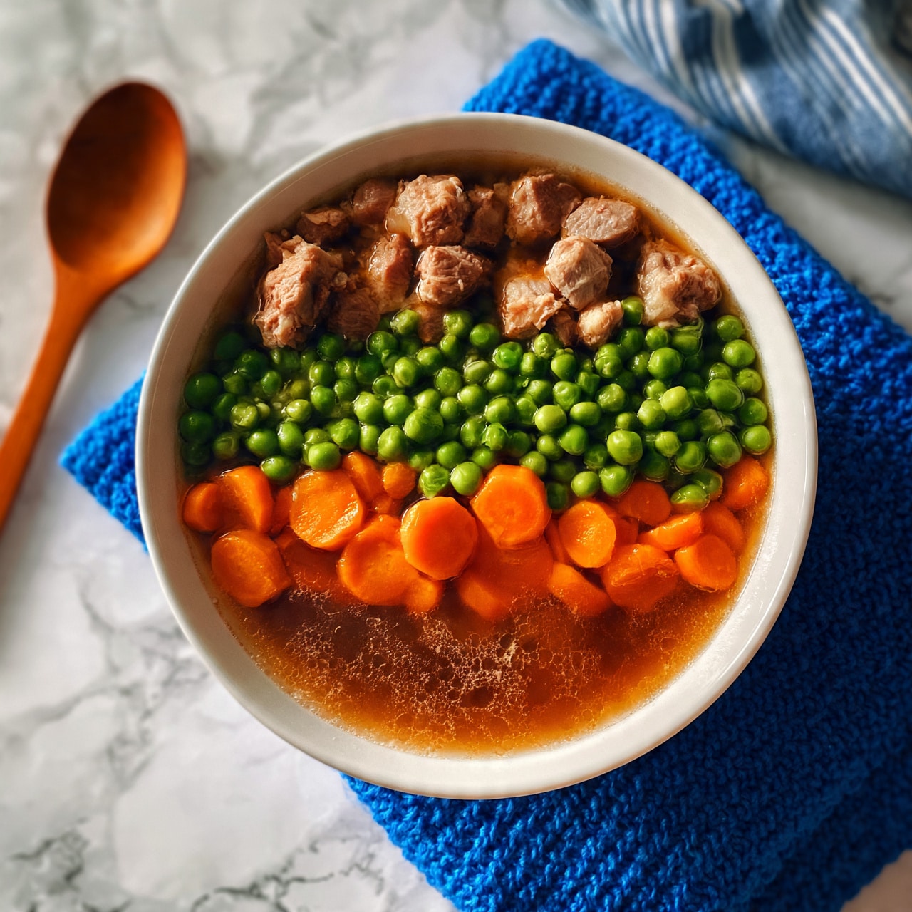 square image A white bowl filled with a colorful stew sits on a white marbled surface. The stew has three layers visible: the bottom layer is brown broth with a slightly shiny texture, the middle layer is a mix of orange sliced carrots and green peas spread evenly, and the top layer shows chunks of light brown meat scattered throughout. The bowl rests on a blue knitted cloth, and there is a wooden spoon placed to the left side on the white marbled surface. A blue and white striped cloth is partially visible in the top right corner. Photo taken with an iphone --ar 4:5 --v 7