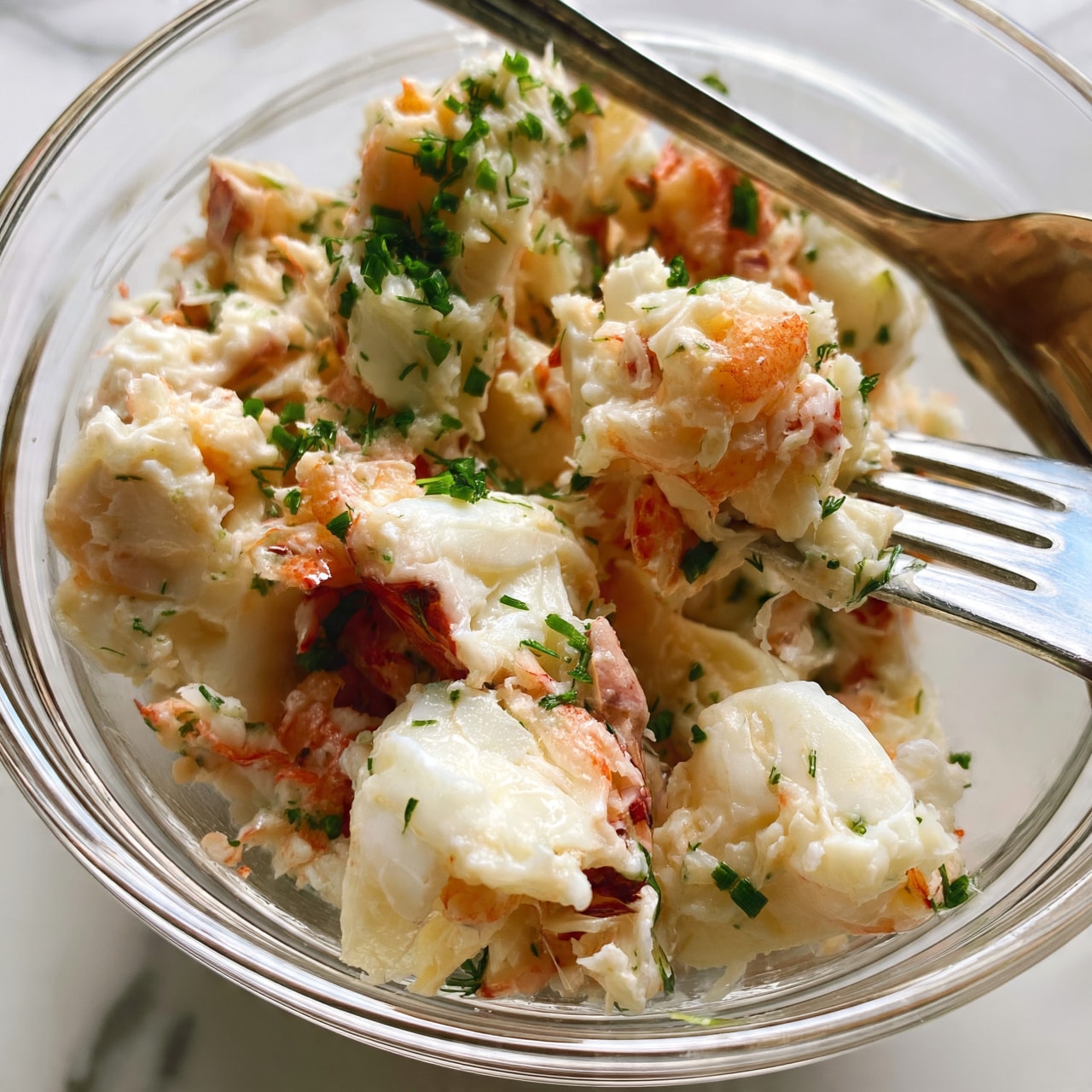 square imag The image shows a close-up of a clear glass bowl filled with pieces of cooked white lobster meat that have light orange and pale reddish parts from the shell. The lobster meat looks soft and has small green herbs, like chopped chives and dill, scattered evenly on top and mixed within. A silver fork is resting inside the bowl, partially holding some of the lobster meat. The bowl is placed on a white marbled surface, and the photo is taken close up to show the texture and details of the lobster meat and herbs. photo taken with an iphone --ar 4:5 --v 7