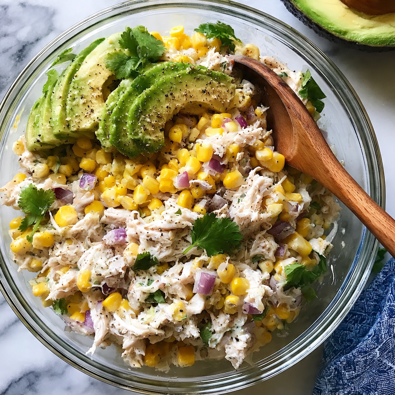 square image A clear glass bowl filled with a mixed salad that has four main layers: shredded white chicken pieces at the base, bright yellow corn kernels scattered throughout, green avocado slices on top, and small chunks of purple onion spread evenly across. The salad is lightly seasoned with black pepper and garnished with fresh green cilantro leaves. To the right inside the bowl, there is a wooden spoon resting. The bowl sits on a white marbled surface with half an avocado visible in the top right corner and a blue patterned cloth in the bottom right corner. Photo taken with an iphone --ar 4:5 --v 7