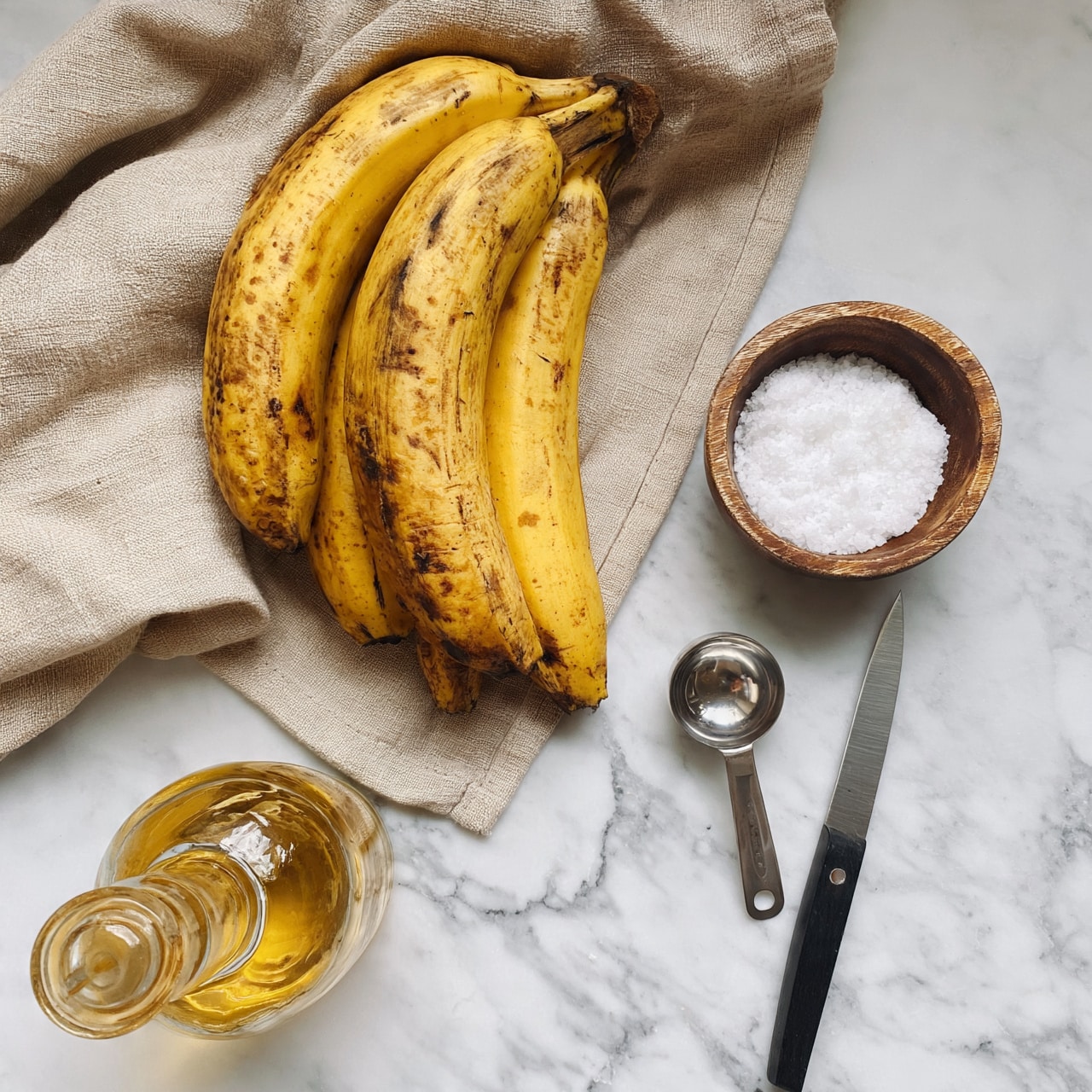 square image A bunch of four ripe yellow plantains with some brown spots rests on a beige cloth draped over a white marbled surface. To the right of the plantains is a small wooden bowl filled with white coarse salt. Below the bowl, a silver measuring spoon and a sharp knife with a black handle are placed side by side on the white marbled surface. On the lower left side of the image, there is a glass bottle filled with a clear golden liquid, likely oil, with a narrow neck and a round stopper. The light is soft, creating a clean and simple composition. photo taken with an iphone --ar 4:5 --v 7